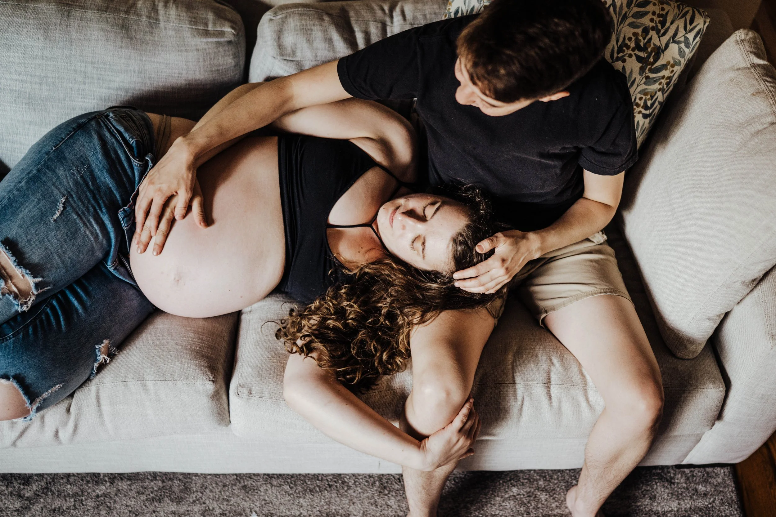 A couple lying on a sofa, embracing, with a pregnant woman resting her head on the man's chest, while the man gently holds her head and stomach.