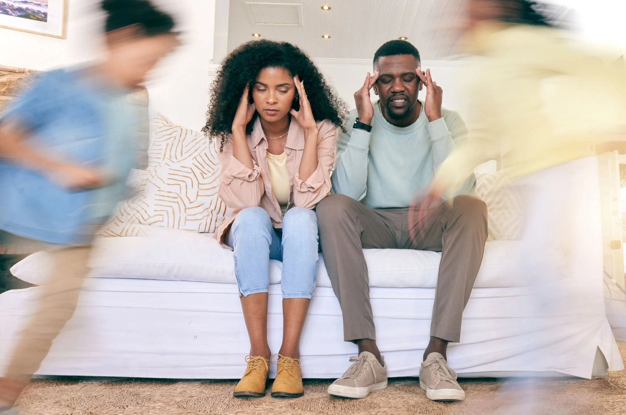 Two people sitting on a white couch with their eyes closed and hands on their temples, while two blurred individuals stand nearby. The woman has curly black hair and wears a beige jacket, and the man has short dark hair and wears a light gray sweatshirt.