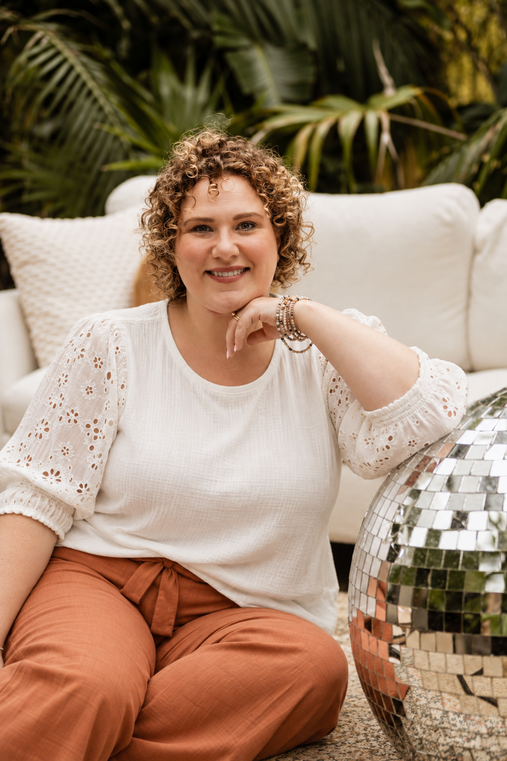 A woman with short curly hair sitting on the floor next to a reflective mosaic table, smiling, with a white sofa and pillows behind her, surrounded by lush green plants.