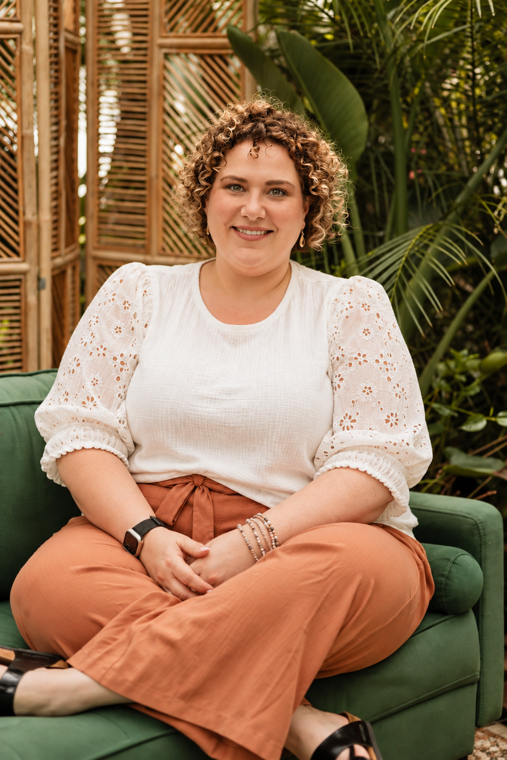 A woman with curly hair smiling, sitting on a green couch outdoors with green plants and a wooden lattice divider in the background. She is wearing a white blouse with eyelet details, rust-colored wide-leg pants, and accessories including earrings, bracelets, and a watch.