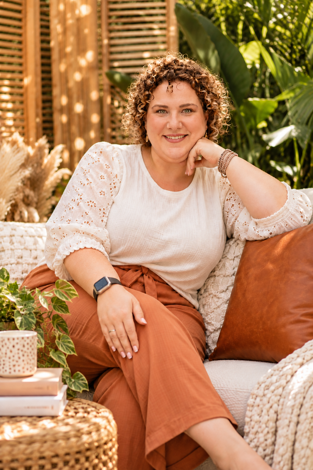 A woman with curly hair and a white blouse sitting on a cozy outdoor couch surrounded by green plants, smiling at the camera.