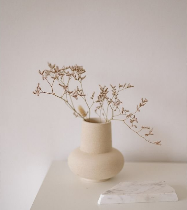 A beige ceramic vase with a rounded base and a narrow neck, containing dried white flowers with small buds, placed on a white surface against a plain light-colored wall.