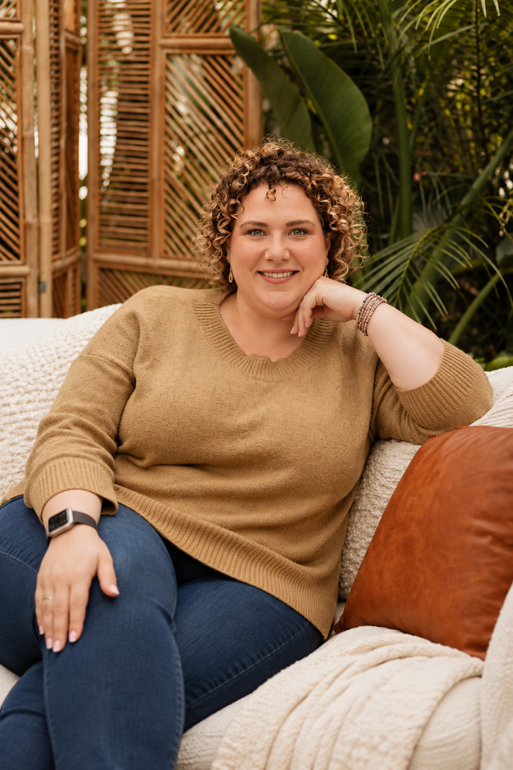 A woman with curly short hair, wearing a tan sweater and blue jeans, sitting on a cream couch with a brown pillow, smiling at the camera, with a background of greenery and bamboo fencing.