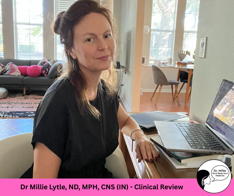 Dr Millie at her desk. A woman with brown hair and a black top sitting at a wooden desk with a laptop and notebooks in a room with large windows and a view of outdoors.