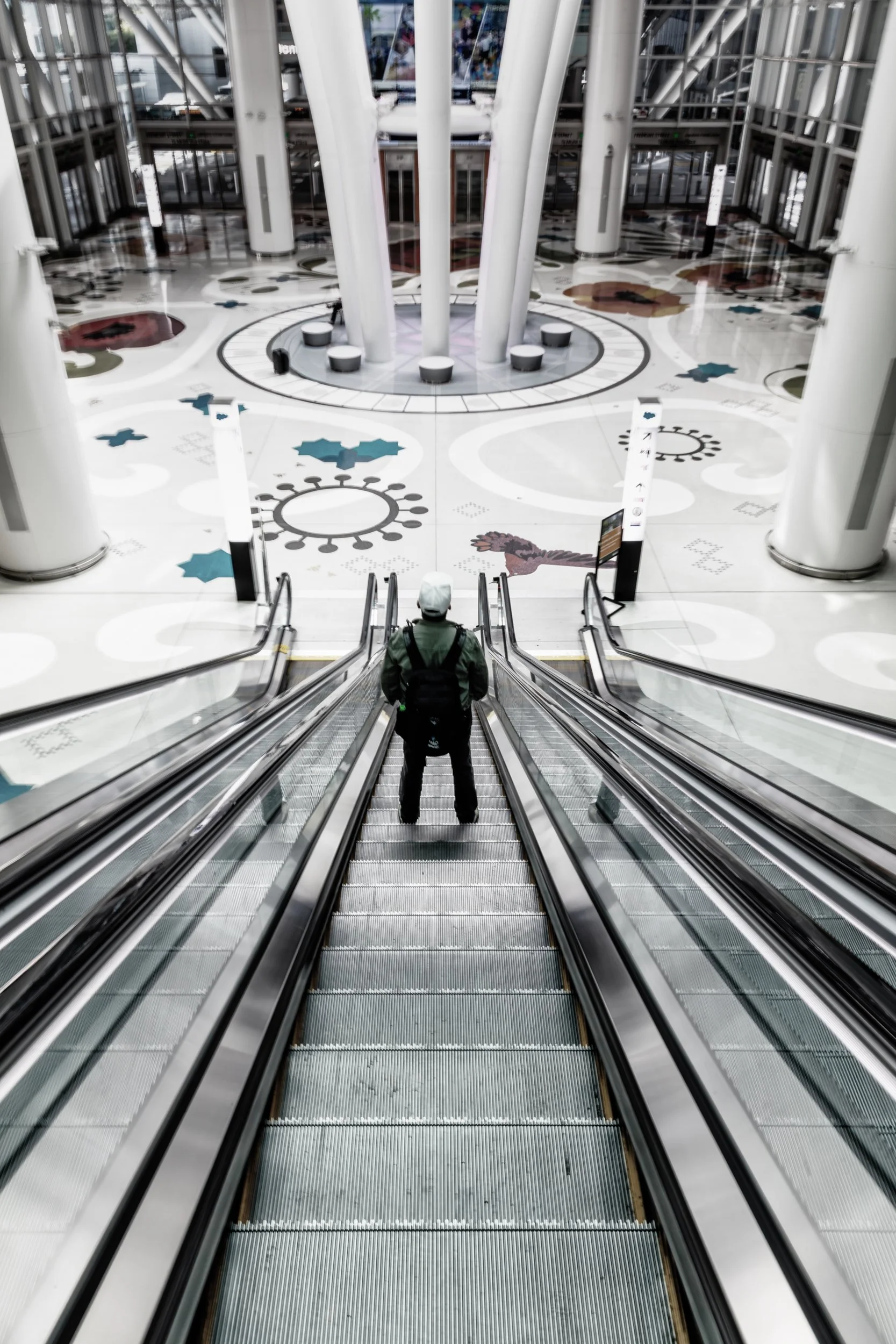 Descent at Salesforce Transit Center

 

  A person descends an escalator inside Salesforce Transit Center in San Francisco. The public interior combines transit infrastructure, tall white columns, reflective surfaces, and patterned flooring. 