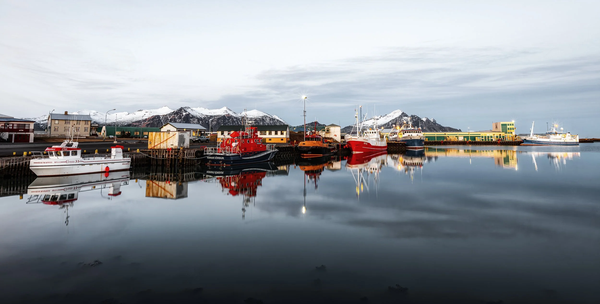 Höfn Harbor Reflections

 

 Fishing boats are moored in the harbor at Höfn, a coastal town in southeast Iceland. Snow-covered mountains, working waterfront buildings, and calm harbor water define the scene. 