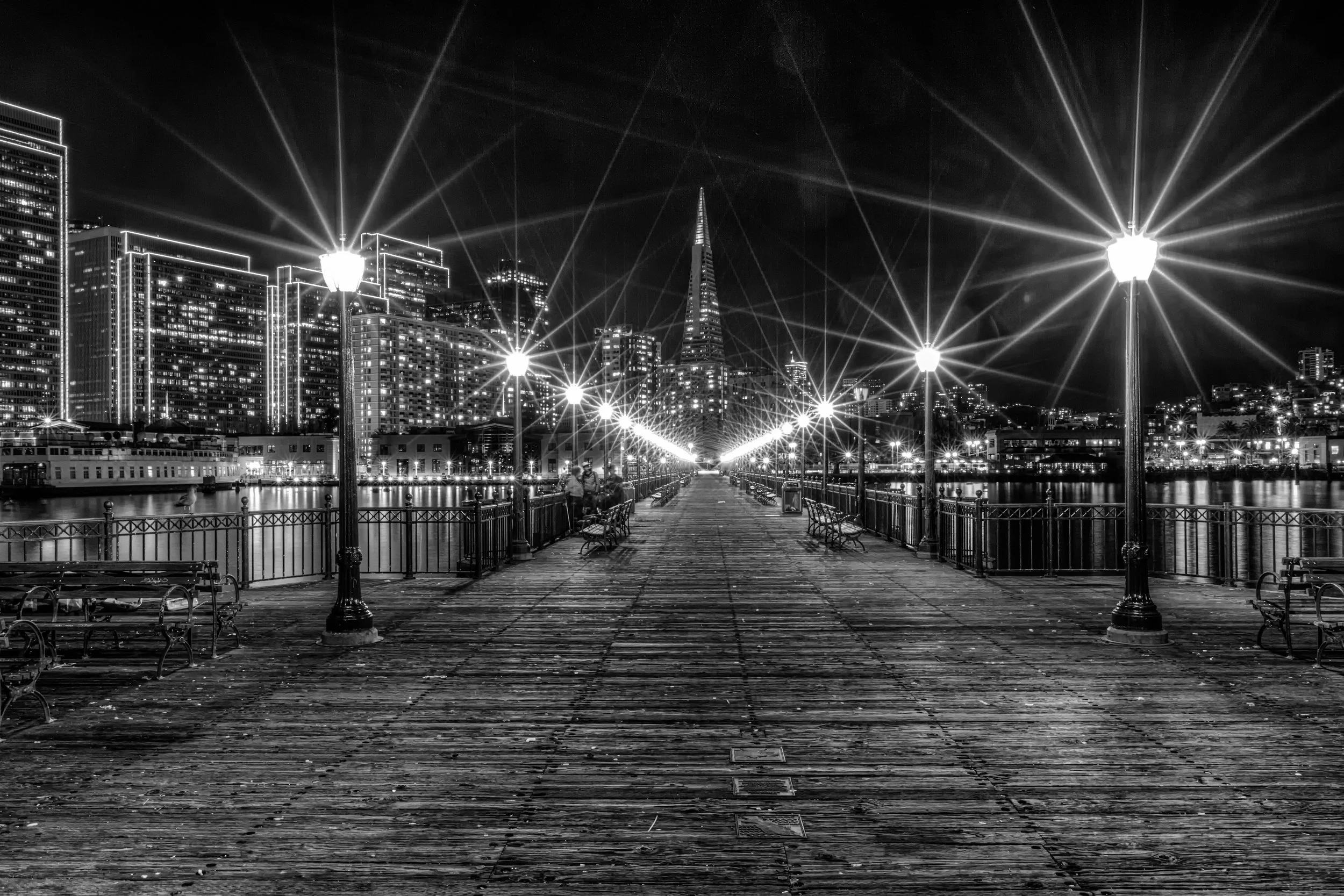 Black-and-white photograph of Pier 7 in San Francisco at night with lights, benches, railings, and the city skyline beyond the wooden deck.