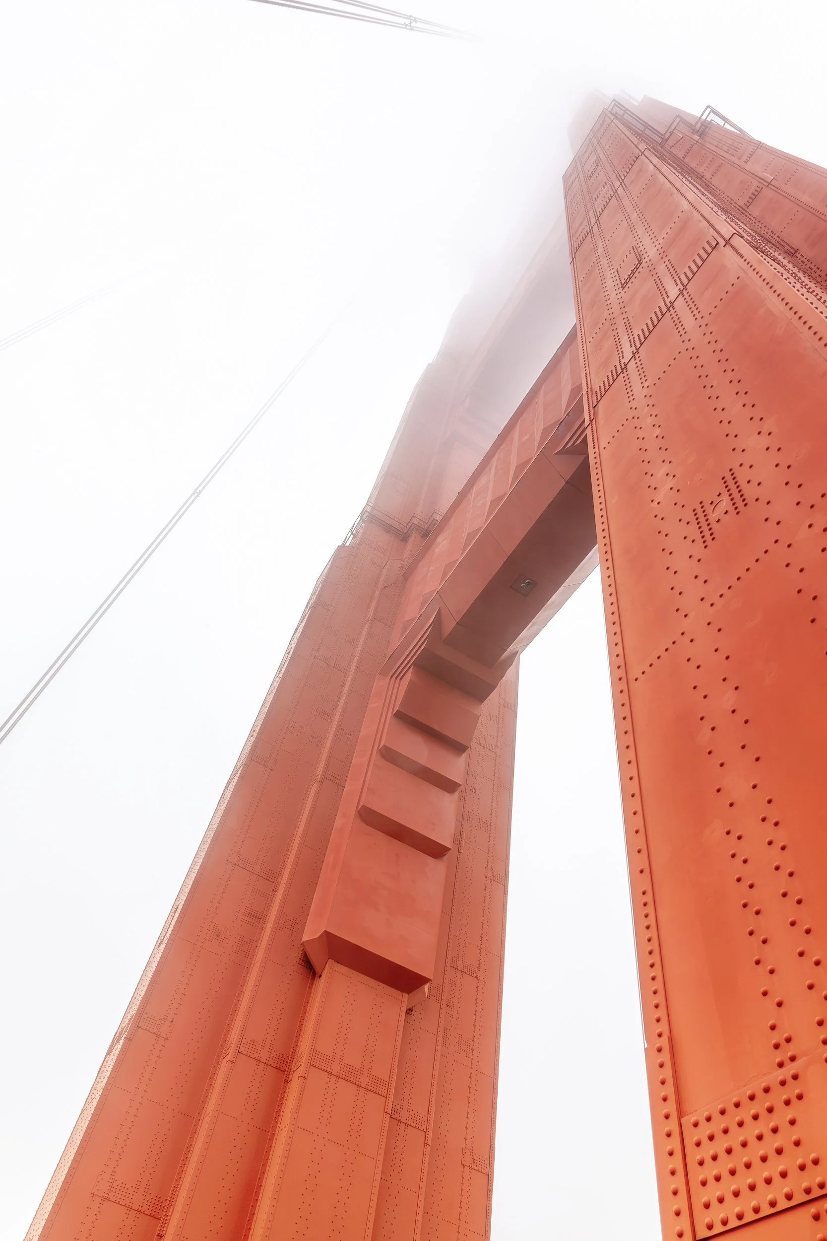 Golden Gate North Tower

 

 The Golden Gate Bridge North Tower rises into dense coastal fog from the bridge’s east sidewalk, with International Orange steelwork and riveted structural panels visible at close range. 