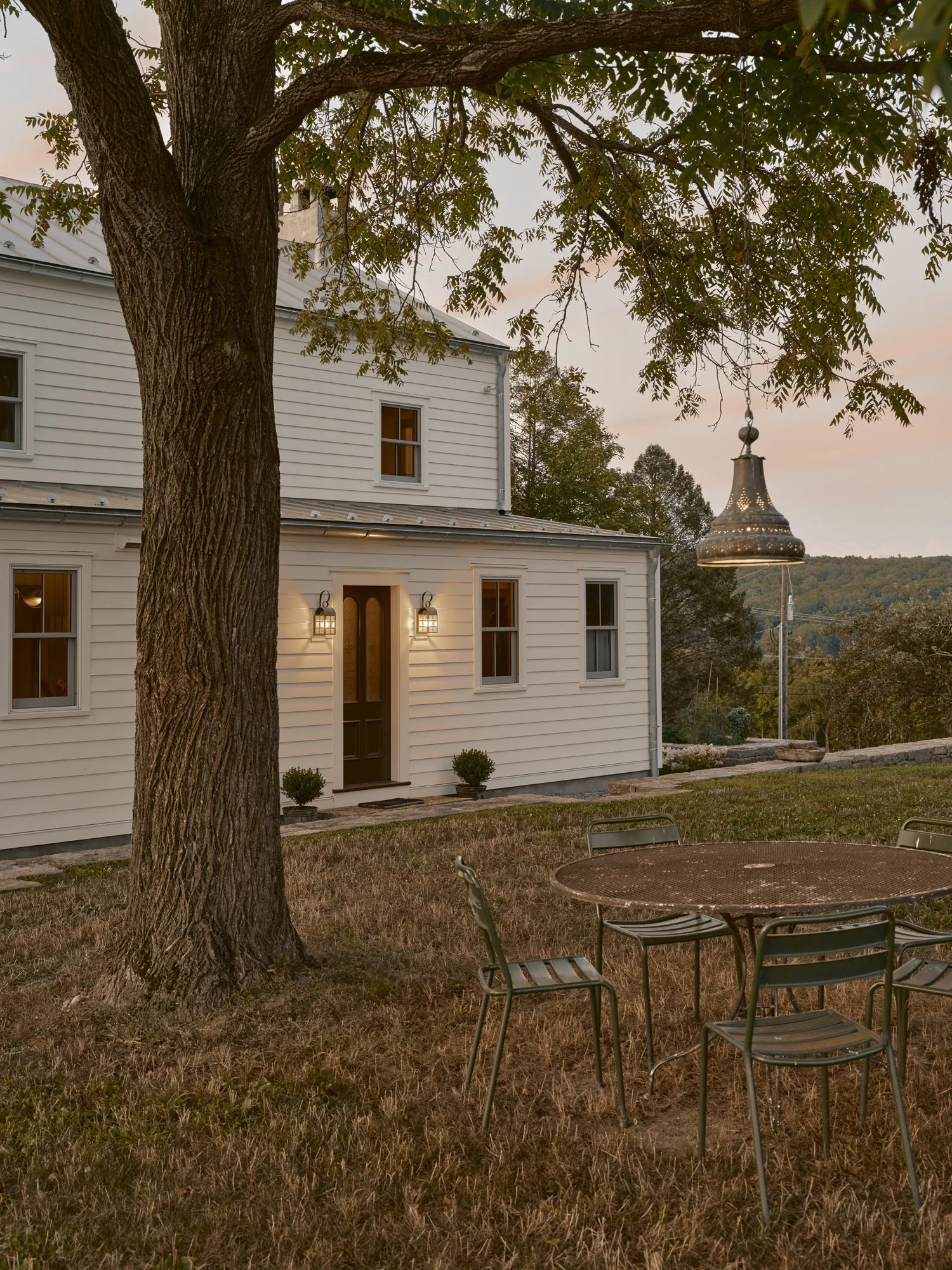 A white house with exterior lights, surrounded by a yard with grass, a large tree, a metal patio table with four chairs, and a hanging lantern near the house during sunset.