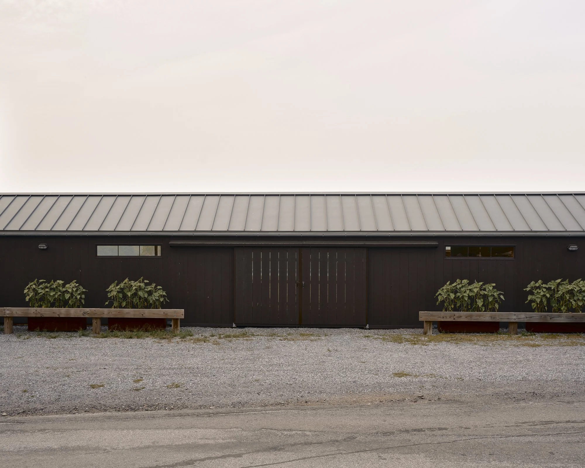 A long, dark-colored building with a metal roof and two narrow windows, surrounded by gravel, with potted plants in front and a pale sky above.