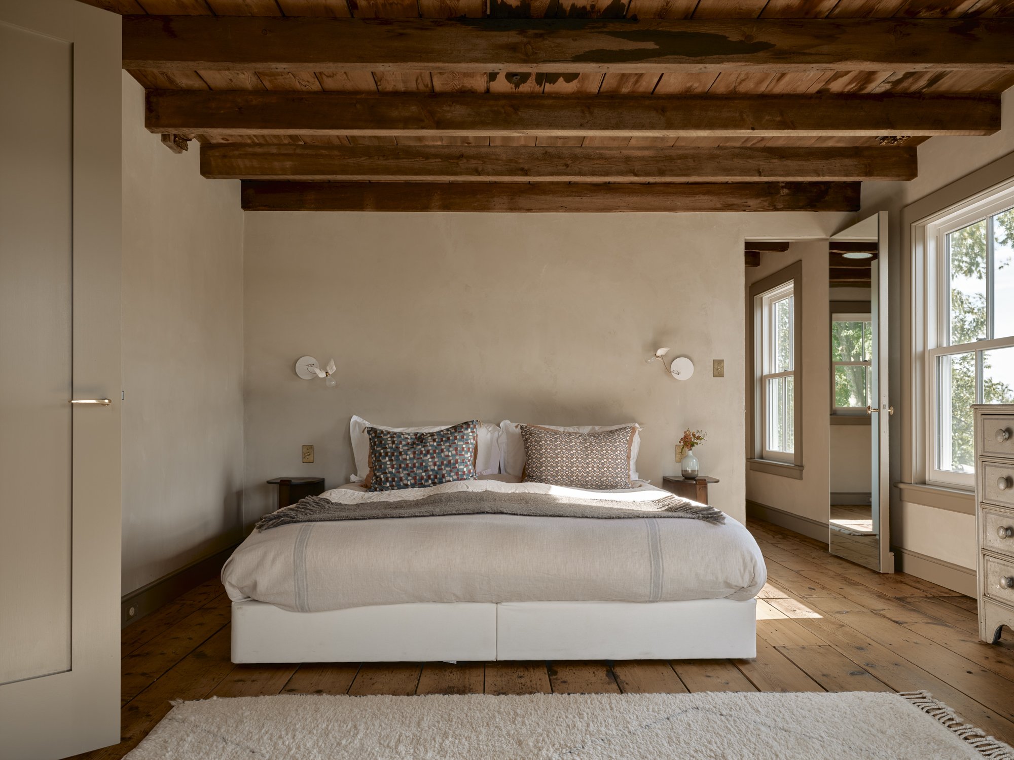 Bedroom with a white bed, two pillows, wooden ceiling beams, large windows, light-colored walls, a small bedside table with a vase, and a dresser.