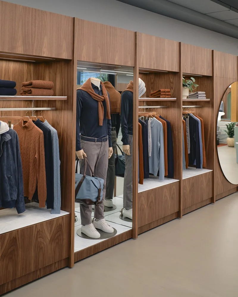 Display of mannequins dressed in casual clothing in a clothing store with wooden shelving and racks holding folded sweaters and hanging shirts.