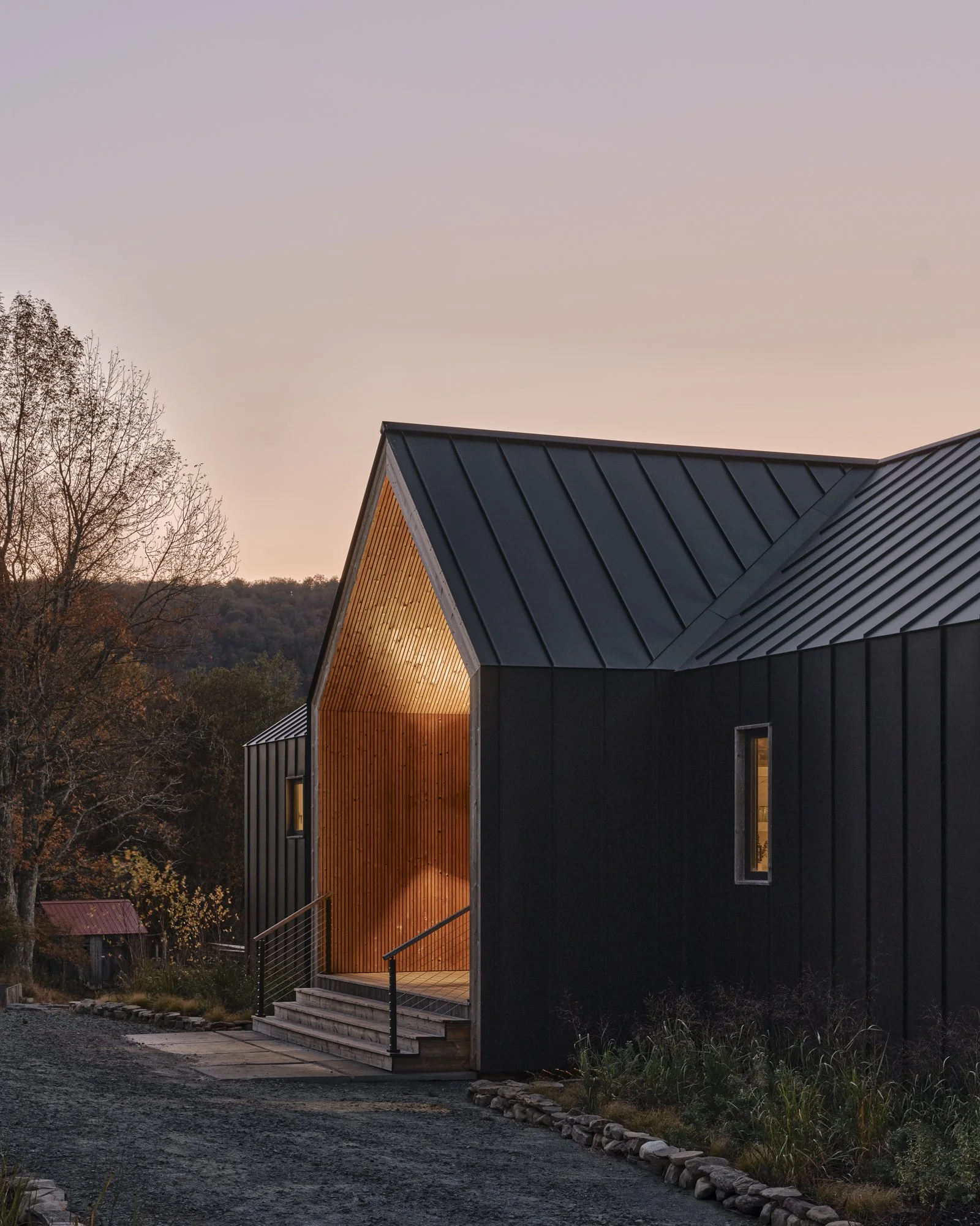 A modern house with black metal exterior and a wooden interior illuminated by warm lighting, set against a backdrop of trees and a pink sky at dusk.