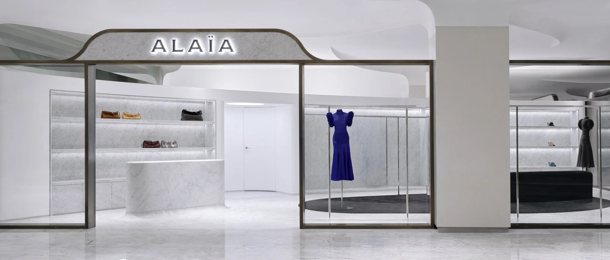 A minimalist clothing boutique with a white marble counter and shelves displaying handbags, a vivid blue dress on a mannequin, and a black dress with shoes on a nearby display.