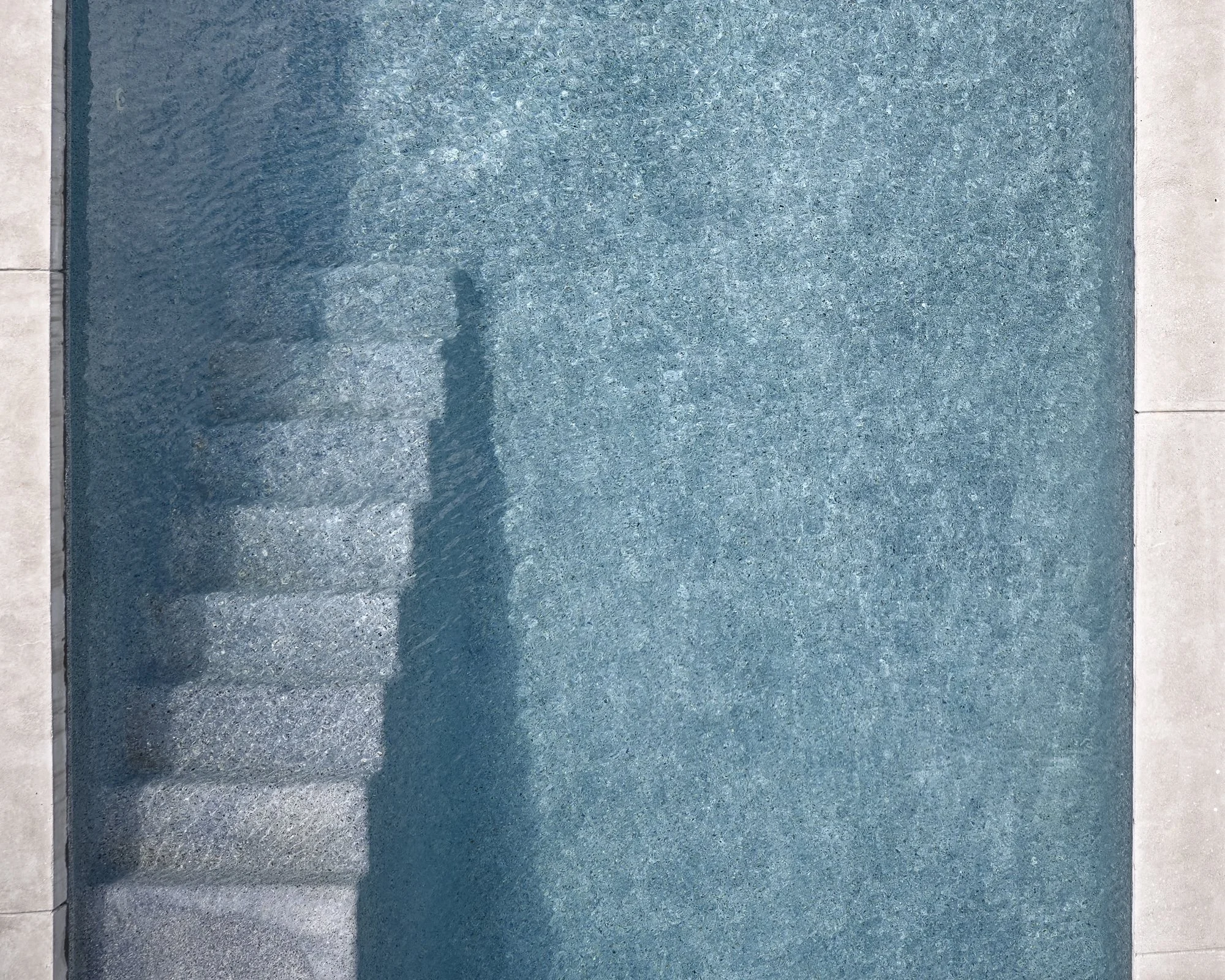 A swimming pool with steps leading into the water, surrounded by light-colored concrete tiles.