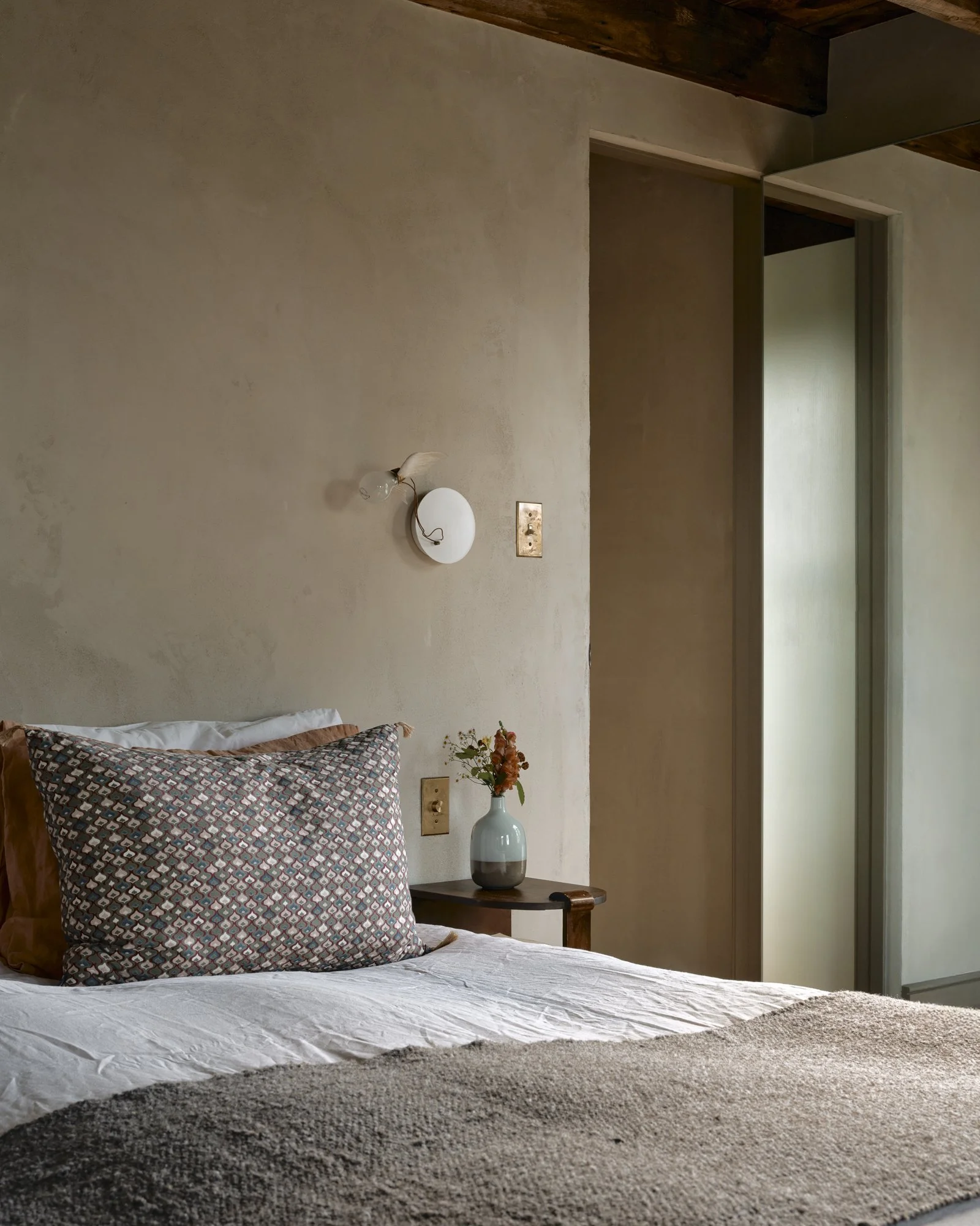 A cozy bedroom featuring a bed with patterned pillows, a beige textured blanket, and a bedside table with a vase of flowers, against a neutral wall with a wall-mounted light fixture and a partially open door.