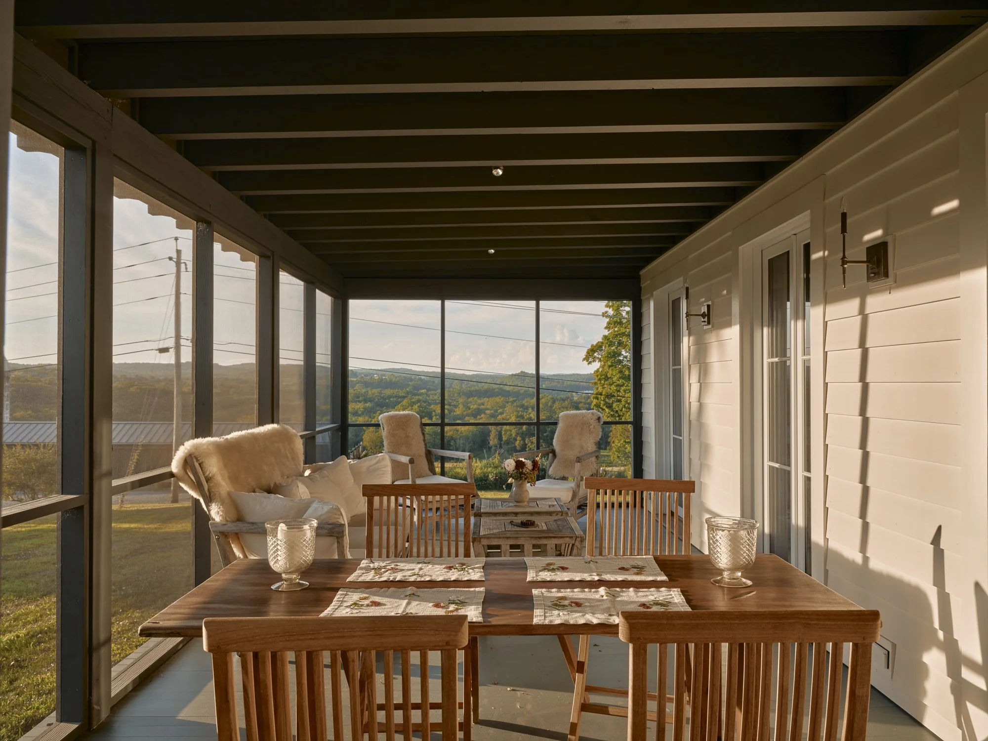 Covered porch with wooden dining table and chairs, cozy seating with cushions and sheepskin throws, overlooking a scenic landscape with hills and trees, during sunset.