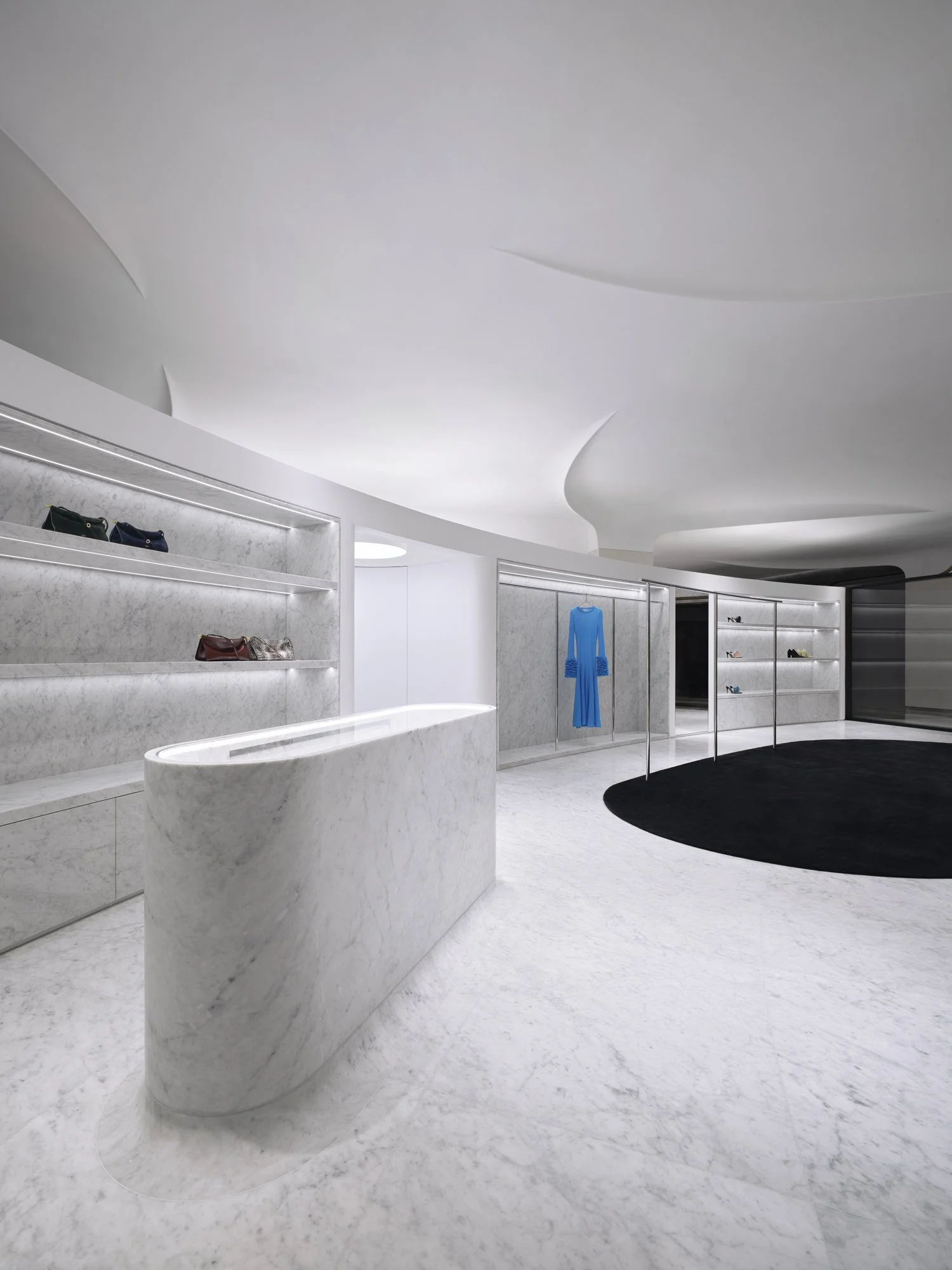 Modern boutique store interior with white marble shelving, curved reception desk, a blue dress on display, and a black circular rug.