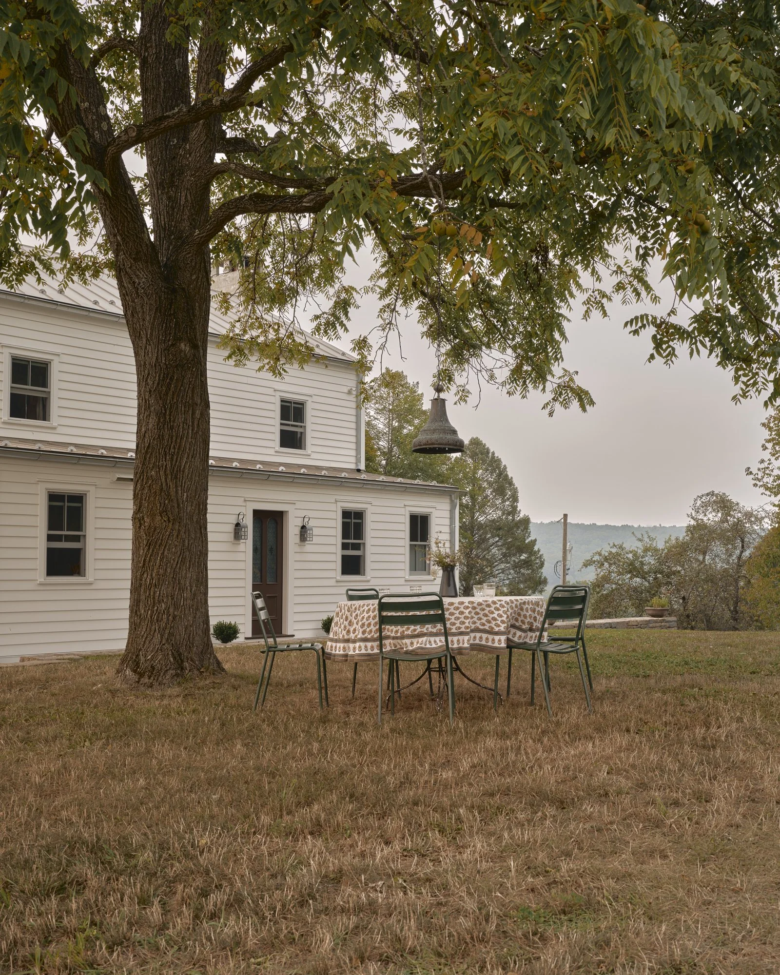 An outdoor scene with a large tree and a white house in the background. There is a round table with a patterned tablecloth and four black chairs on a grassy lawn. A bell hangs from one of the tree's branches.