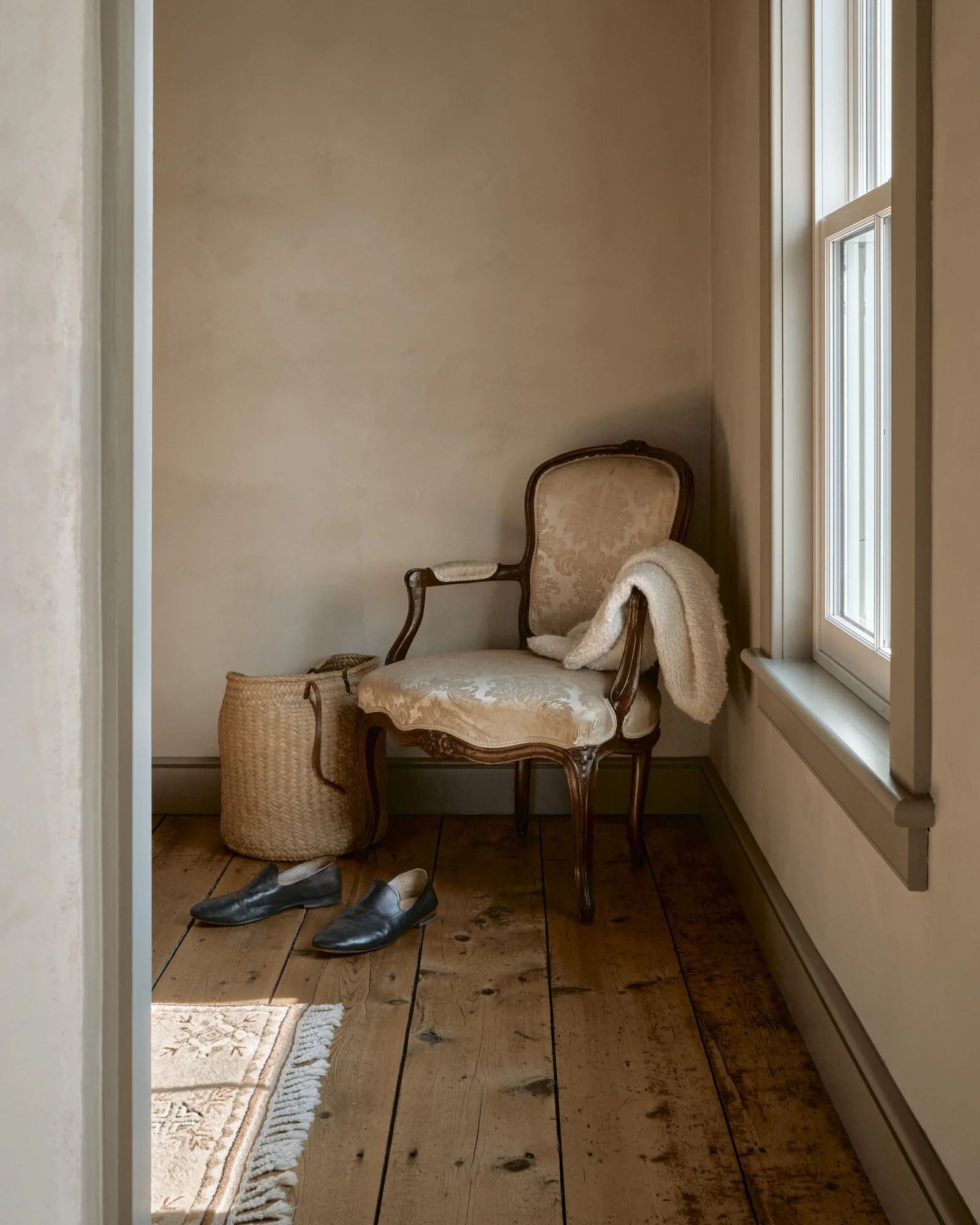 A wooden chair with a beige and gold patterned cushion and backrest, a white blanket draped over one armrest, a woven basket on the floor, and a pair of black shoes nearby, in a room with wooden floors and a window.
