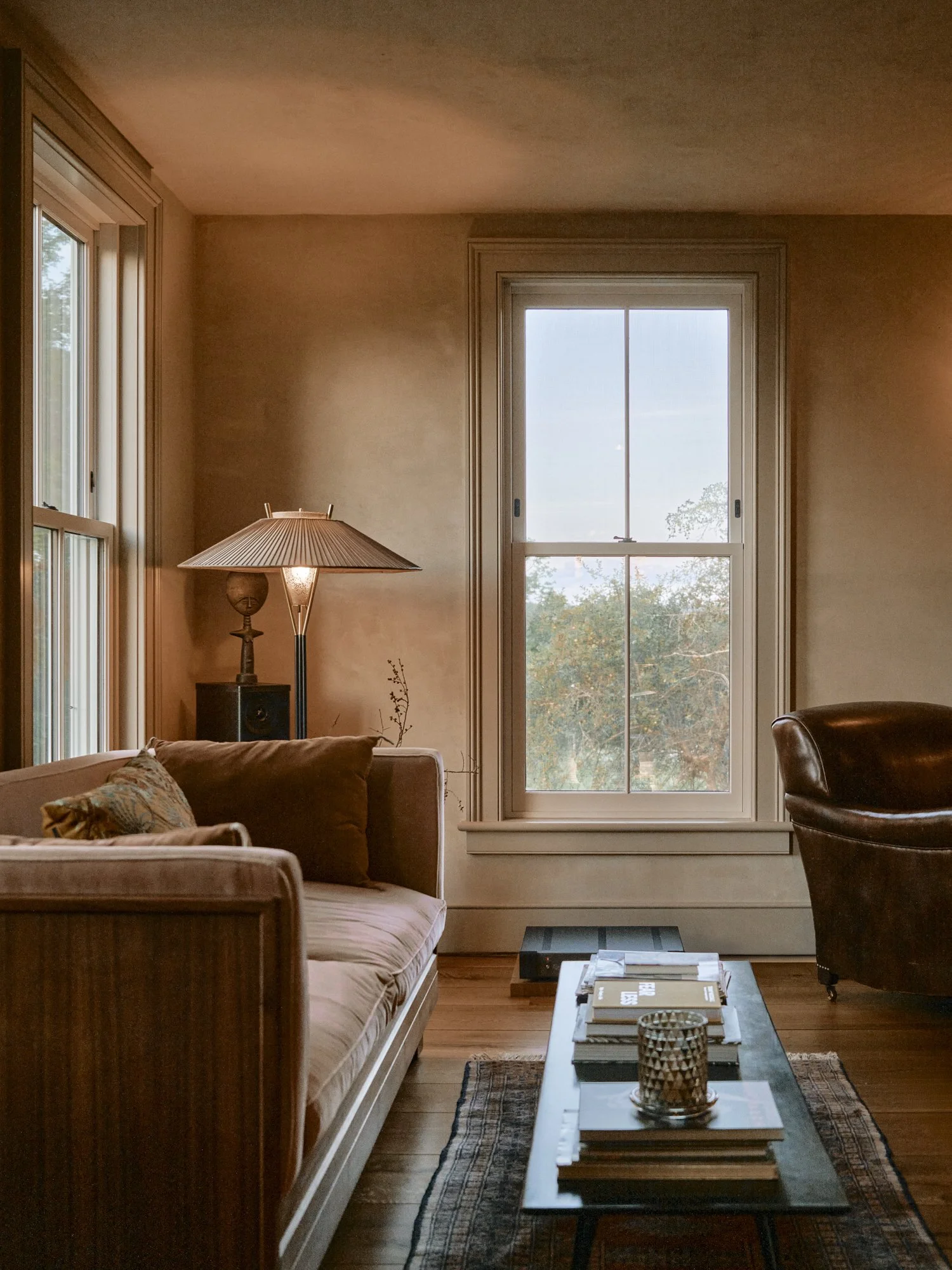 A cozy living room with a wooden sofa, a coffee table with books and a candle holder, a tall window with a view of trees outside, a desk lamp, and a leather armchair.