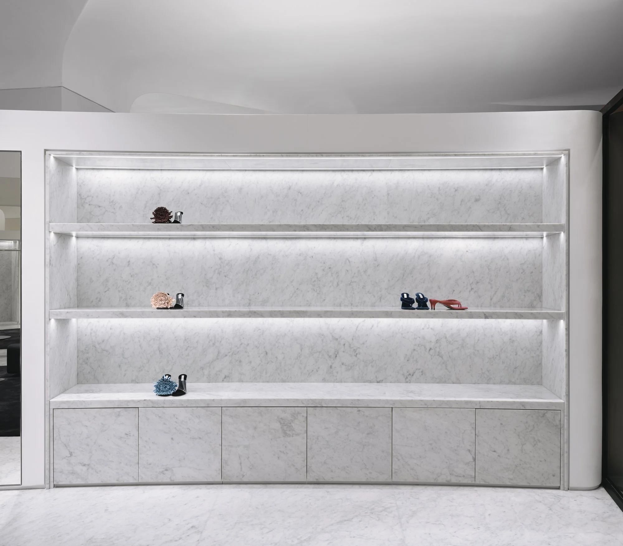 Display of four pairs of high-heeled shoes on a minimalist white marble wall-mounted shelf with built-in lighting.