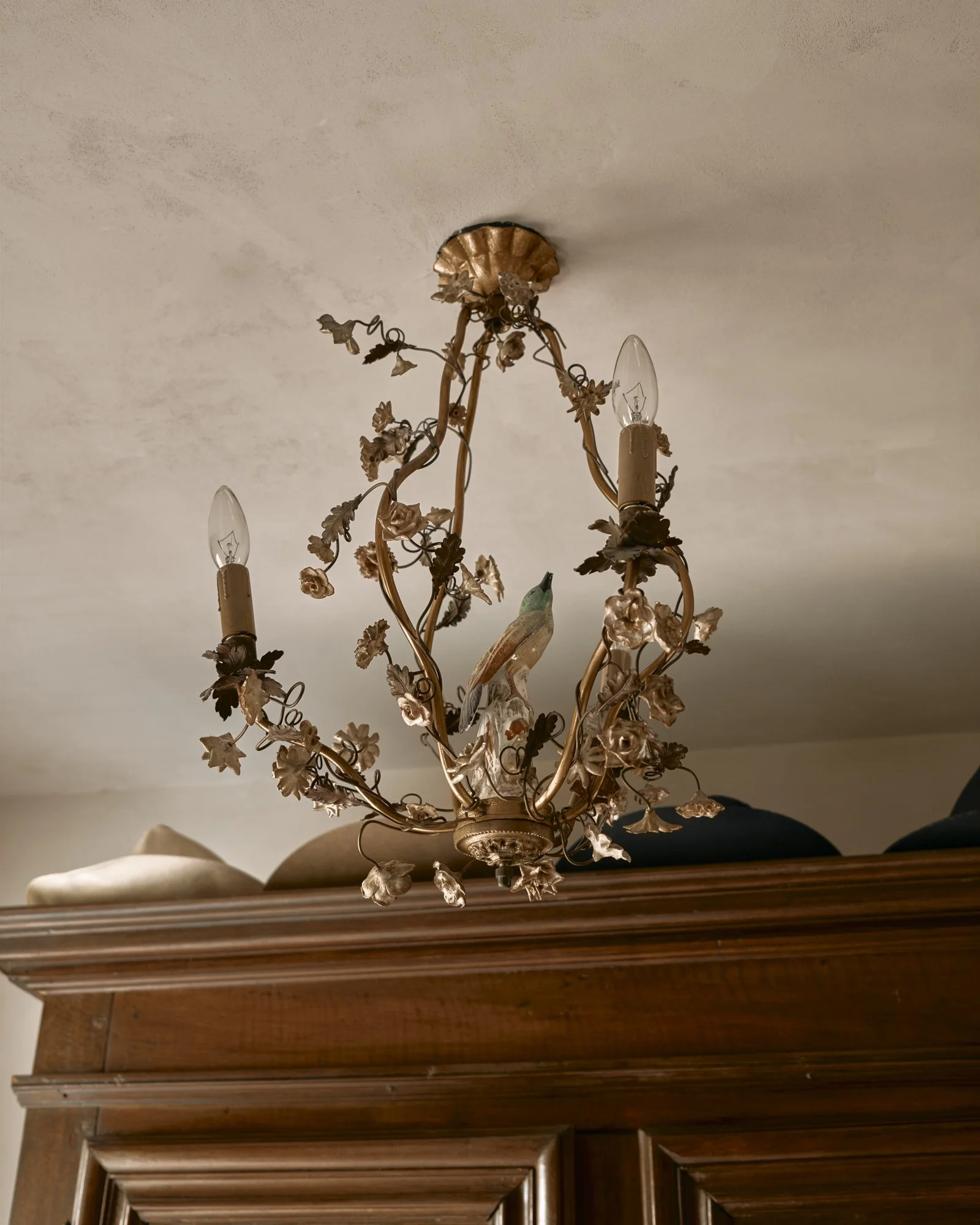 Ornate chandelier with floral details and a bird figurine, mounted on a ceiling above a wooden cabinet.