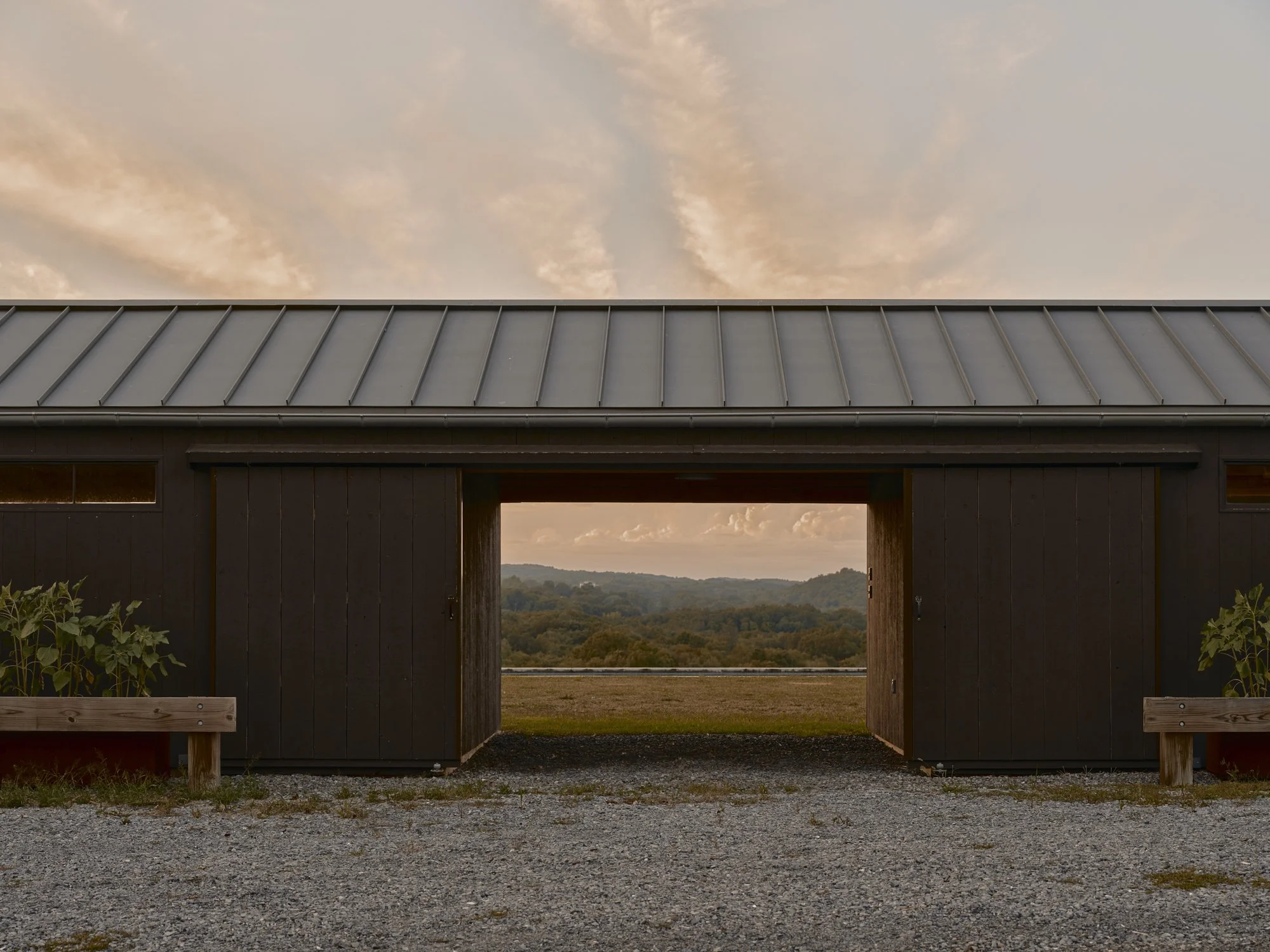 A dark wooden barn with a metal roof and open doors, showing a view of hills and trees in the distance during sunset.