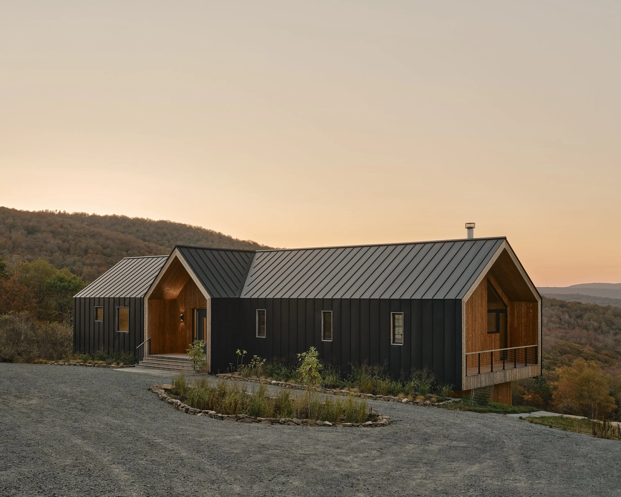 Modern black and wood house on a hill during sunset with a mountain backdrop and gravel driveway