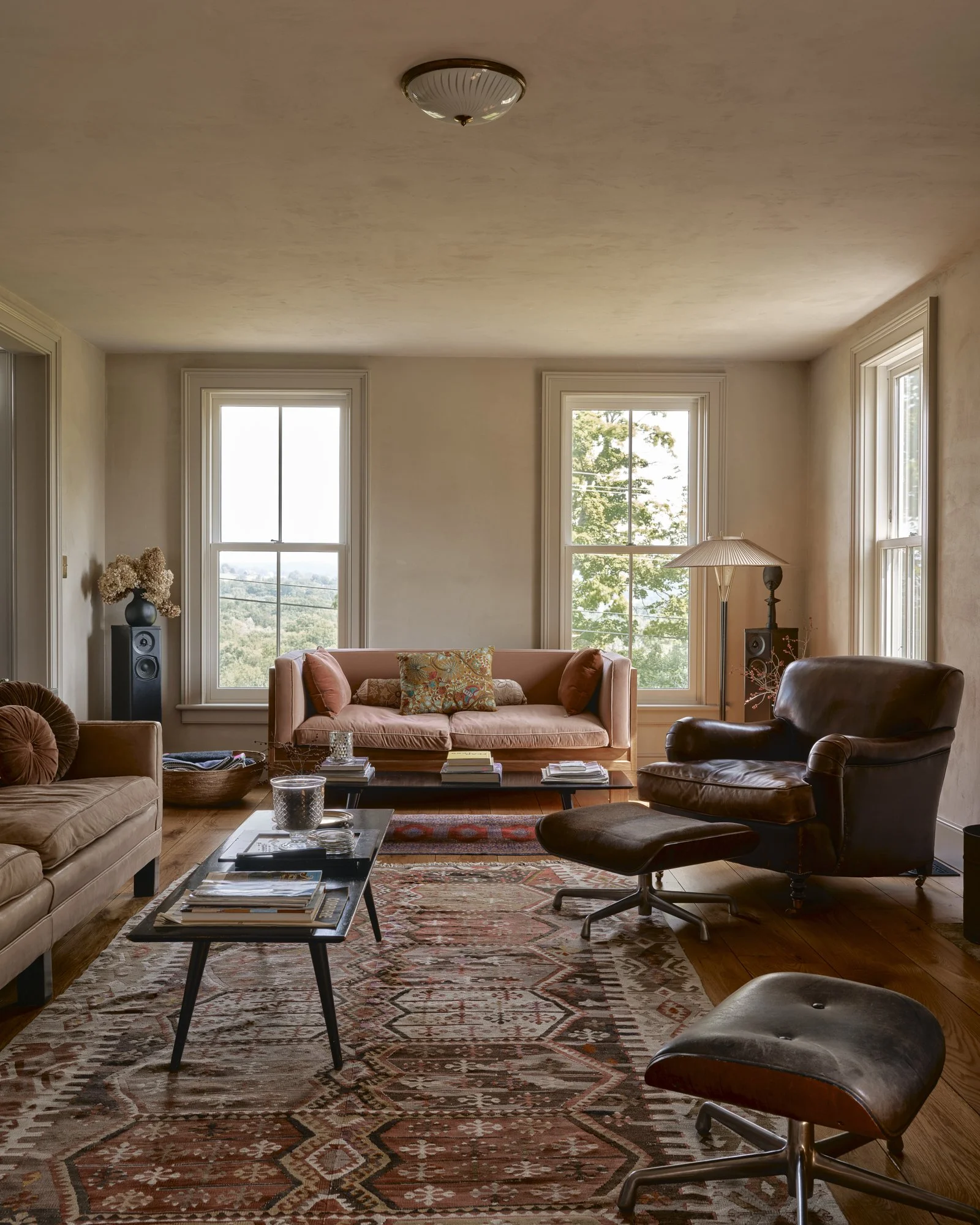 Living room with three large windows, a pink sofa with pillows, a leather armchair with ottoman, a coffee table with books, and a patterned rug.