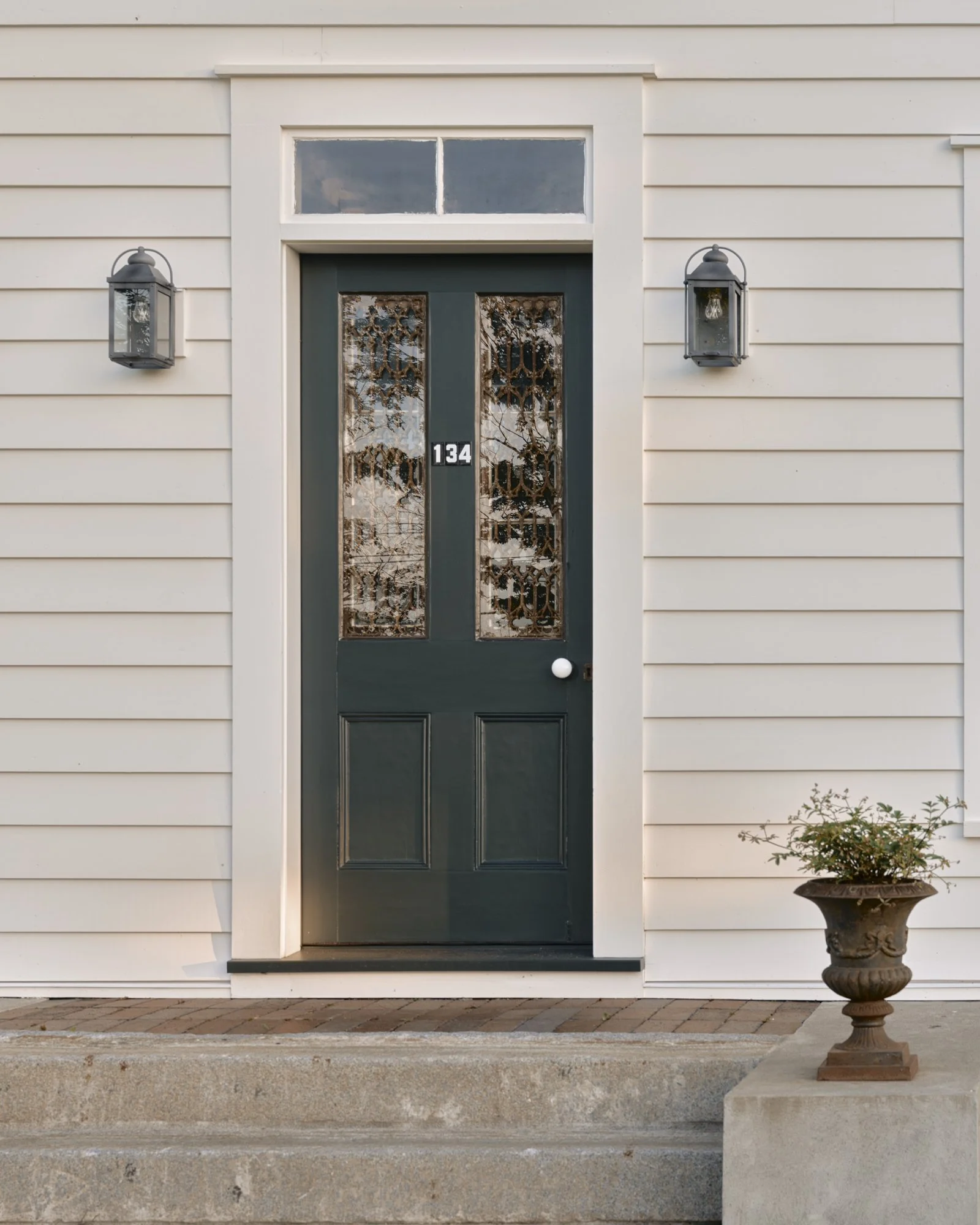 A front door with the number 134, surrounded by white clapboard siding, with two wall-mounted lanterns and a ceramic planter containing a small bush on a concrete step.