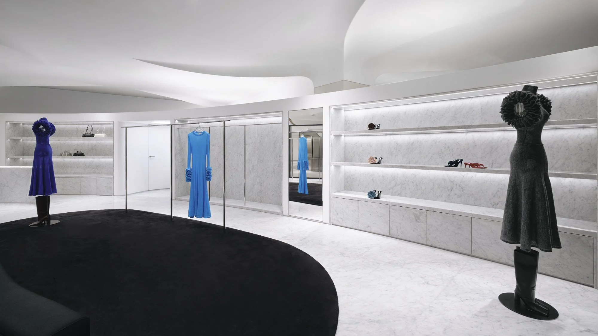 Interior of a luxury clothing and shoe store with white marble walls and floors, displaying mannequins dressed in blue and black dresses, and shelves with various shoes and handbags.
