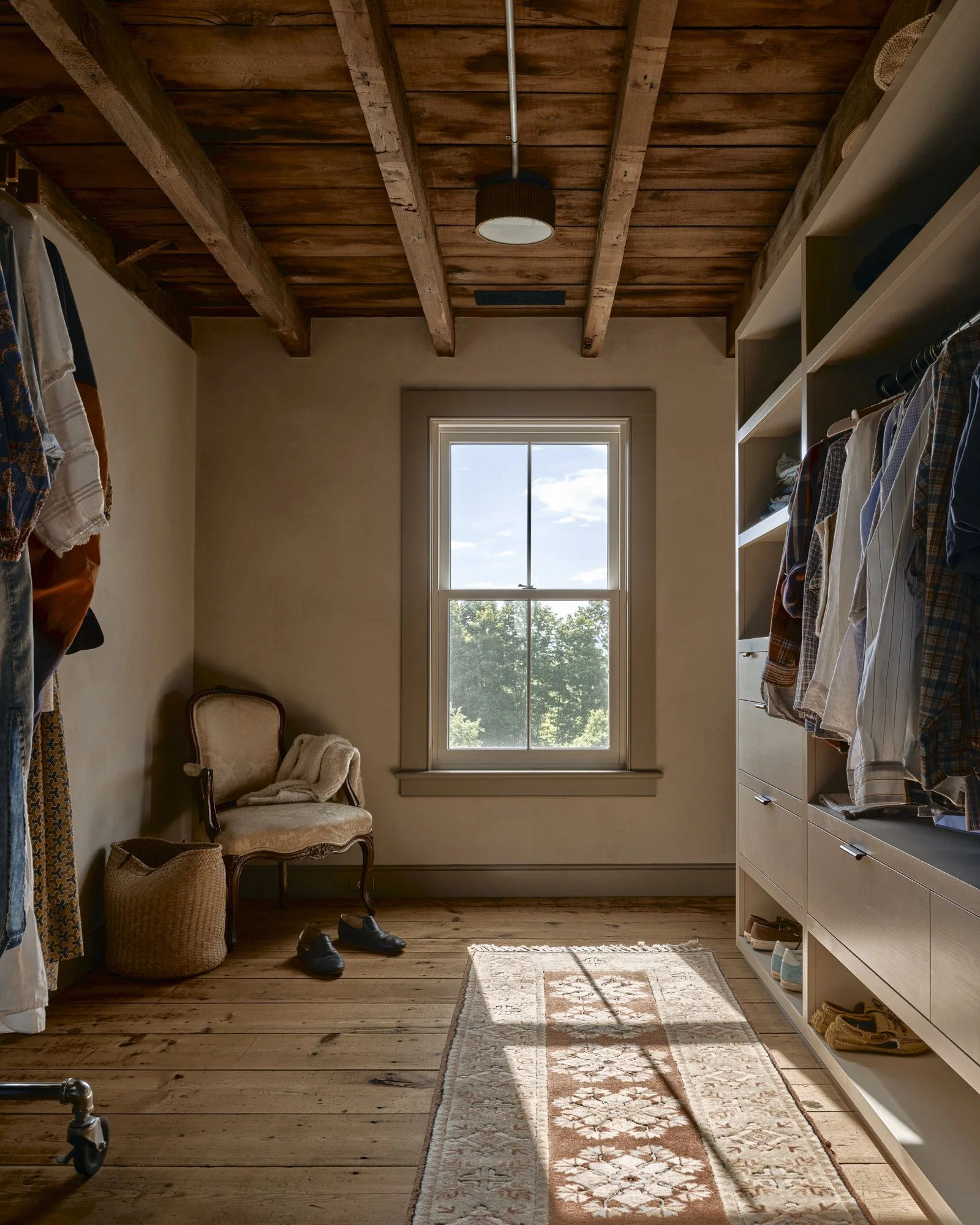 A walk-in closet with wooden floors, a window with a view of trees, and built-in shelves filled with clothing and shoes. There is a vintage armchair, a basket, and a pair of shoes on the floor. Sunlight streams through the window onto a patterned rug