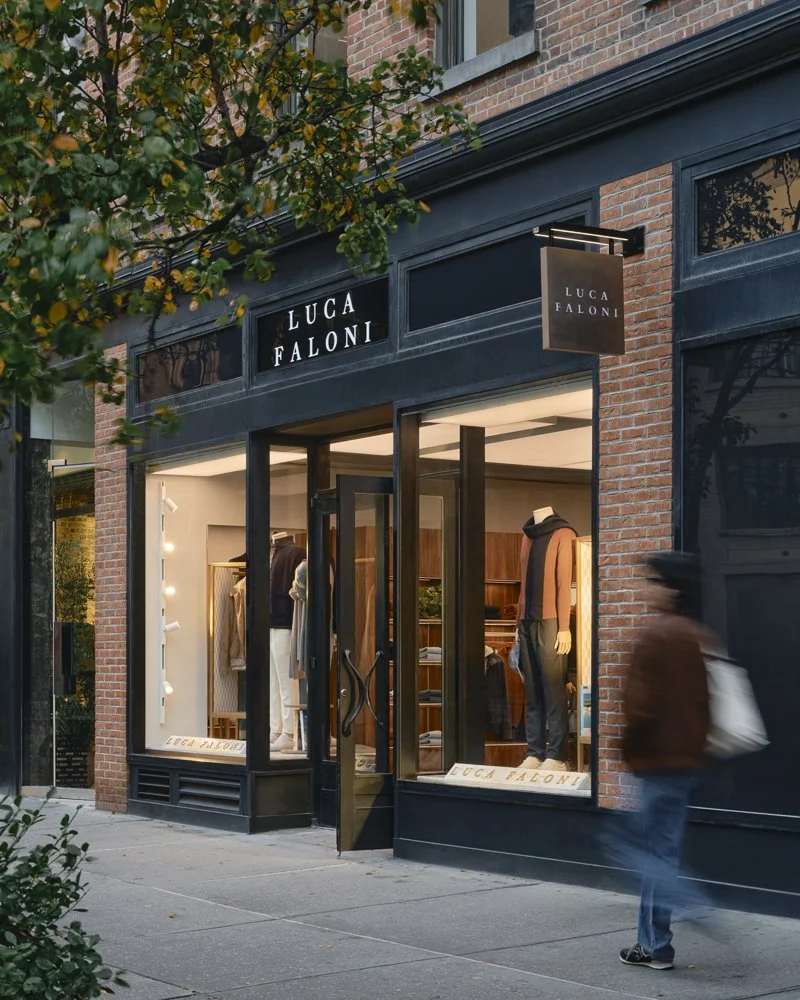 Storefront of LUCA FALONI clothing boutique with mannequins displaying outfits inside, brick and black exterior, signboard and sidewalk, person walking past.