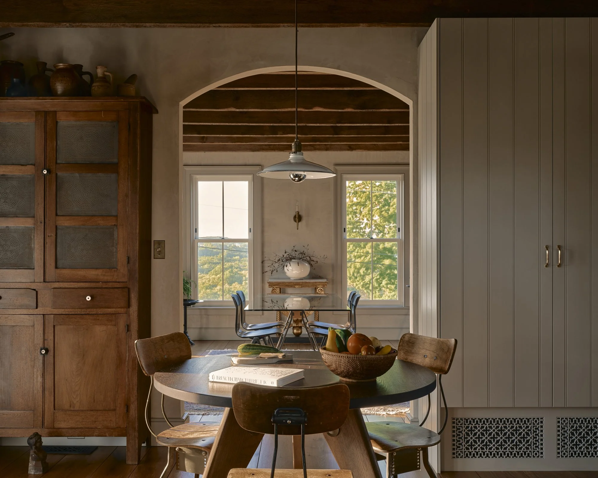 Cozy dining area with wooden and gray cabinets, a round table with chairs, a fruit bowl, and windows with a view of trees.