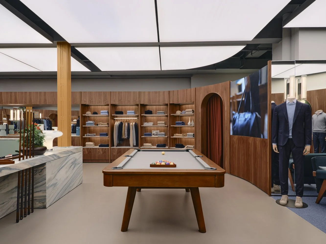 Interior of a clothing store featuring a pool table, wooden shelves with folded clothes, suits on mannequins, and a large TV screen.
