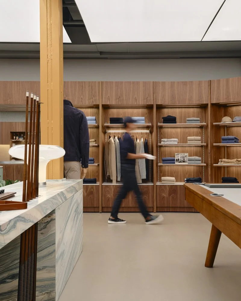 Inside a clothing store featuring wooden shelves with folded clothes and hanging garments, a marble counter with decorative items, and two blurred people browsing.