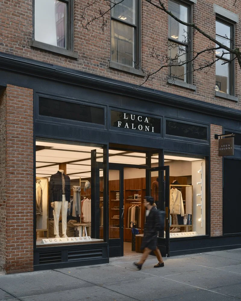 The storefront of a clothing store named 'Luca Faloni' with large display windows showcasing mannequins dressed in stylish apparel. The store is situated in a brick building and a person is walking past on the sidewalk.