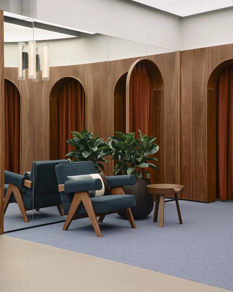 A modern interior with wooden wall panels, arched alcoves with curtains, a mirror, hanging light fixtures, a large potted plant, a blue armchair, a matching loveseat, a small wooden side table, and a gray carpet.