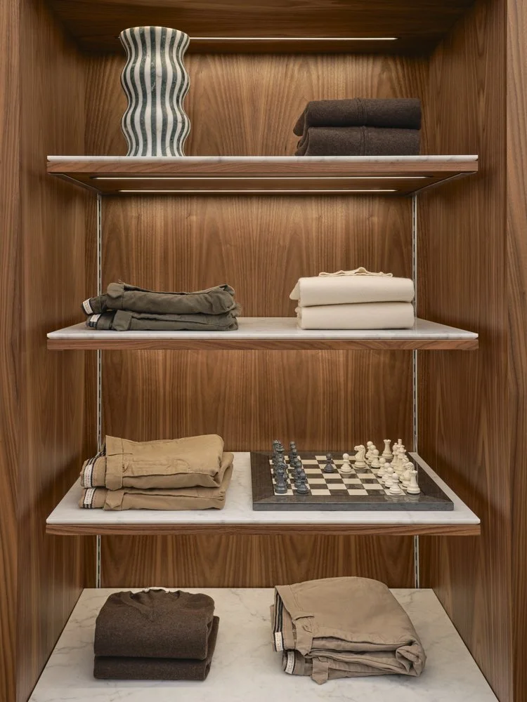 A wooden display shelf with four levels, holding folded pants, a vase, and a chess set.