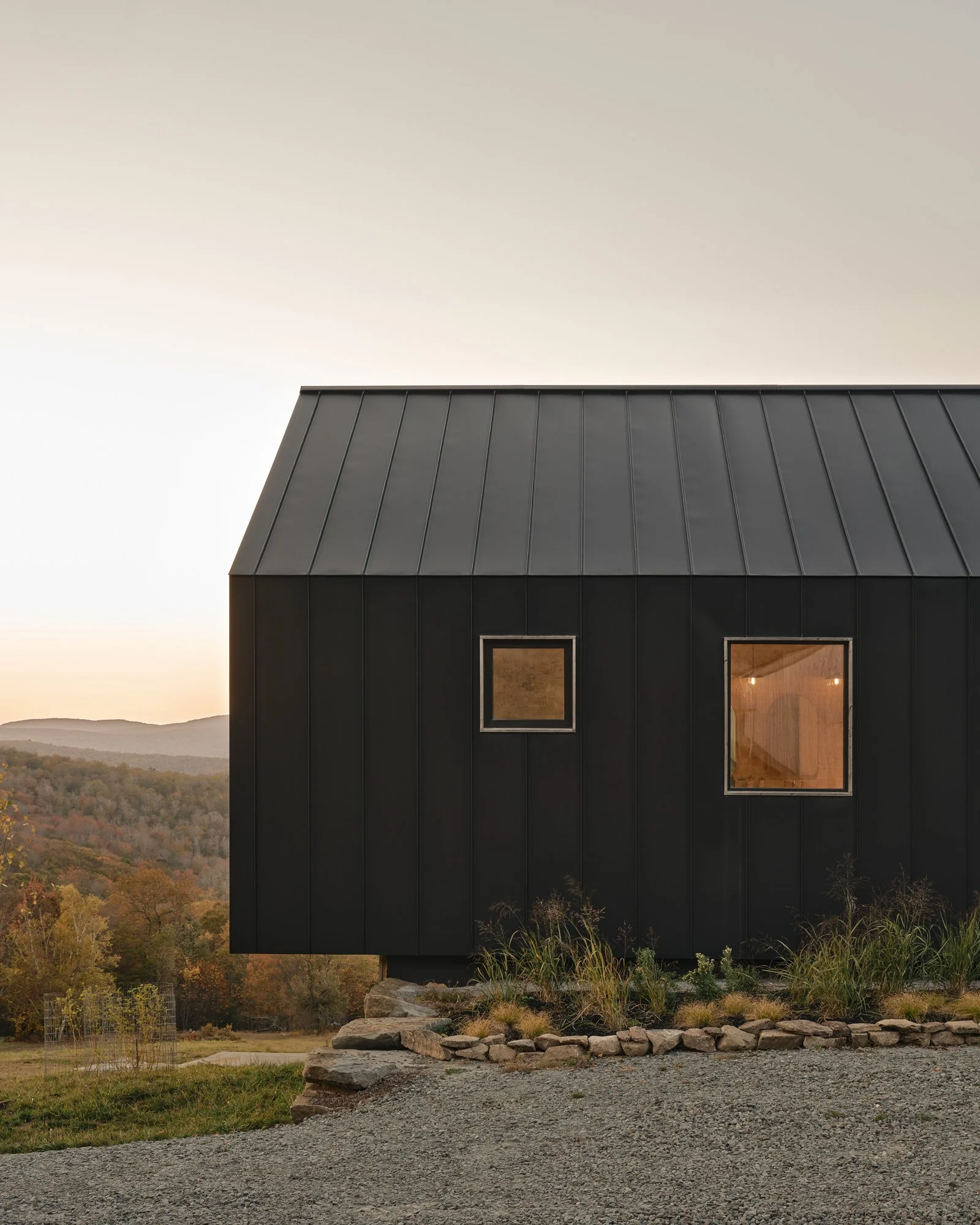 A black house with metal roof and two windows, set against a landscape with trees and hills at sunset.