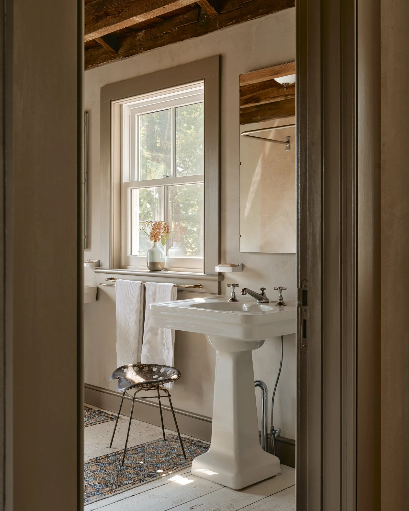 A vintage-style bathroom with a pedestal sink, a mirror, a window with sunlight, a small chair, a vase with pink flowers, and a towel hanging on a brass rail.