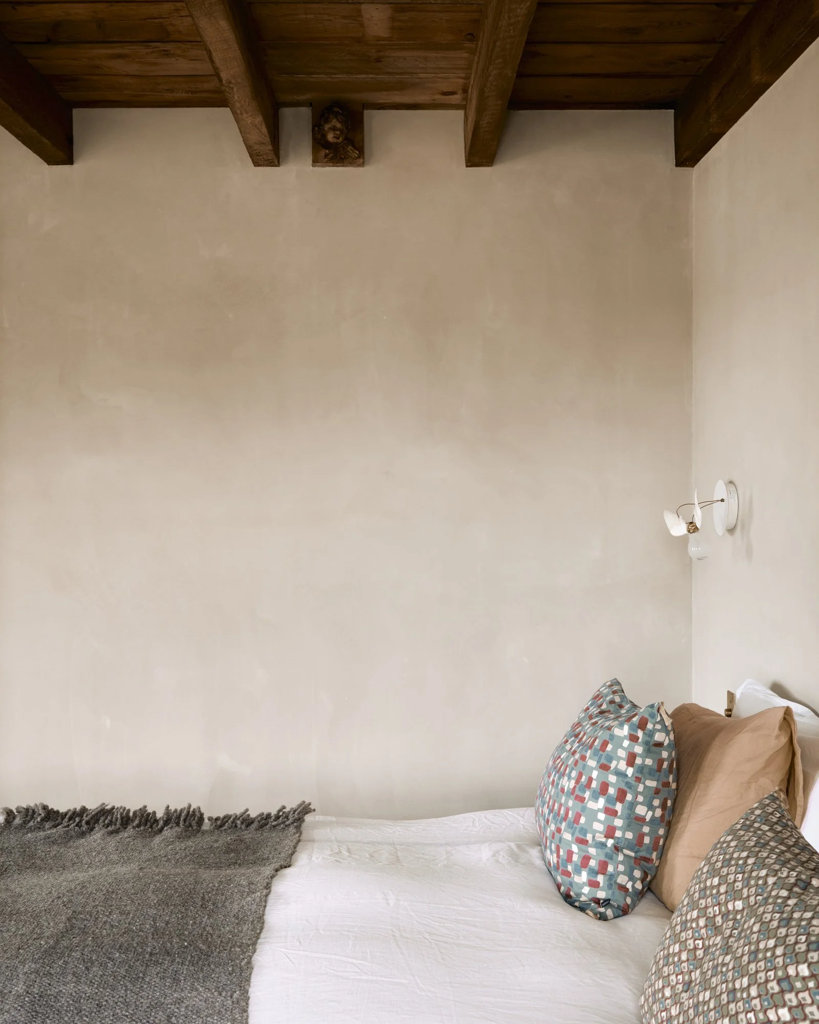 A bedroom with a beige wall, wooden ceiling beams, a white bed with patterned pillows, a brown blanket, and a wall-mounted light fixture.