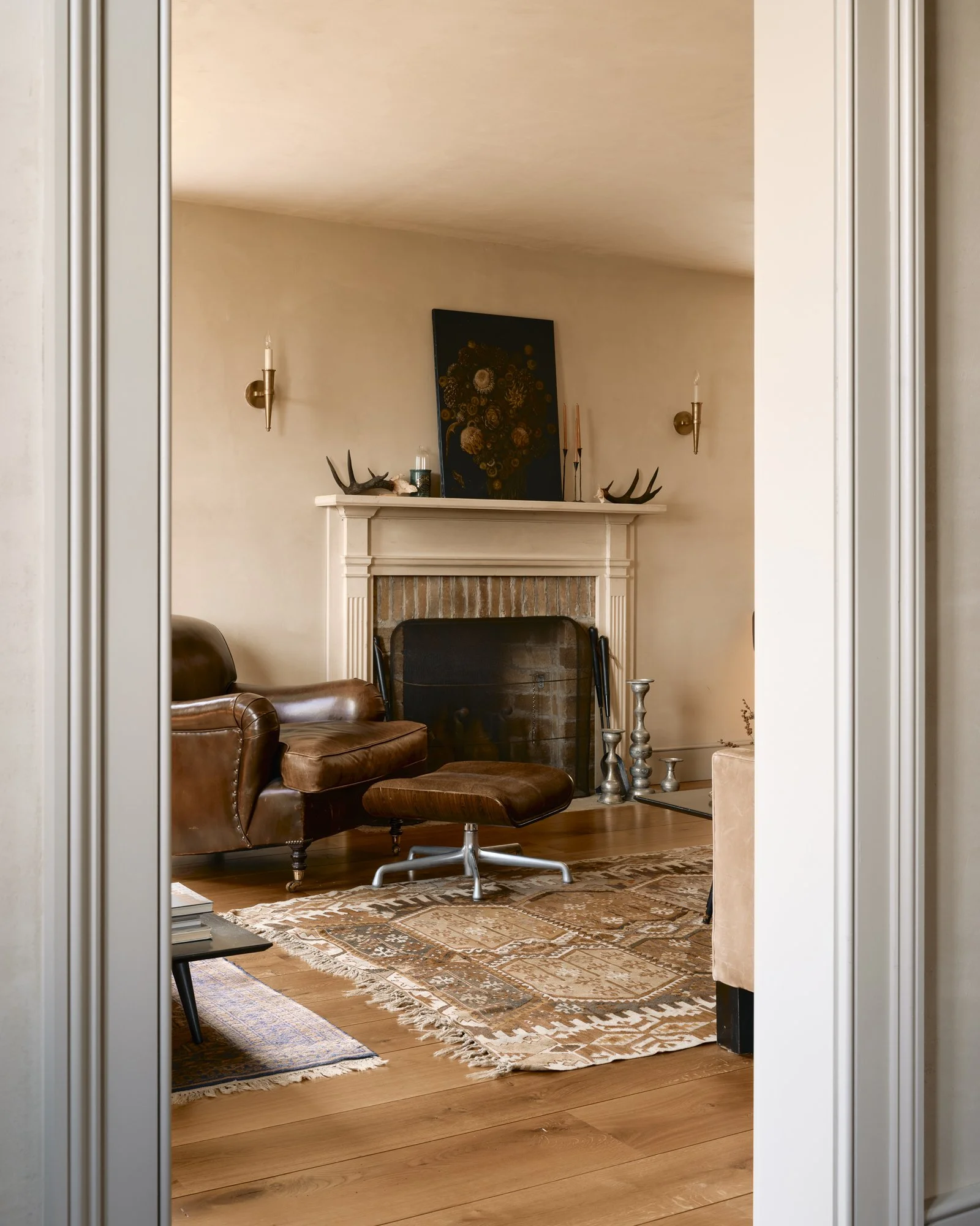 Living room with a leather armchair and matching ottoman, fireplace with decorative items on the mantle, wall-mounted candleholders, a patterned rug, and a framed piece of artwork above the fireplace.