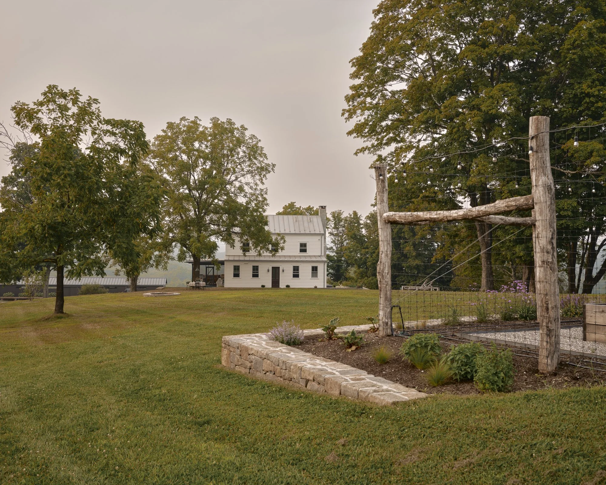A white two-story house in the distance with a grey metal roof surrounded by trees. In the foreground, there is a garden bed with various plants enclosed by a stone border, and a rustic wooden trellis. The lawn is lush and well-maintained, and the sk