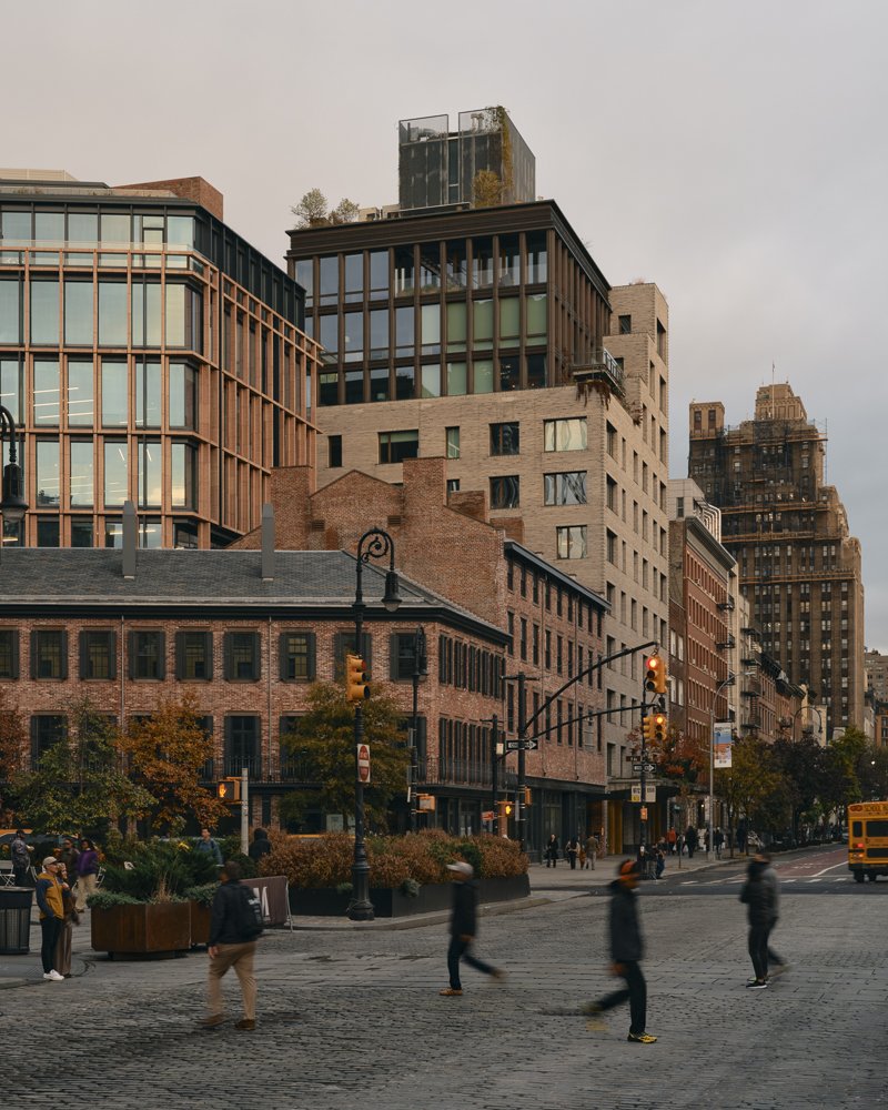 City street scene with pedestrians crossing at traffic lights, brick and glass buildings, and a yellow bus in the distance, under an overcast sky.