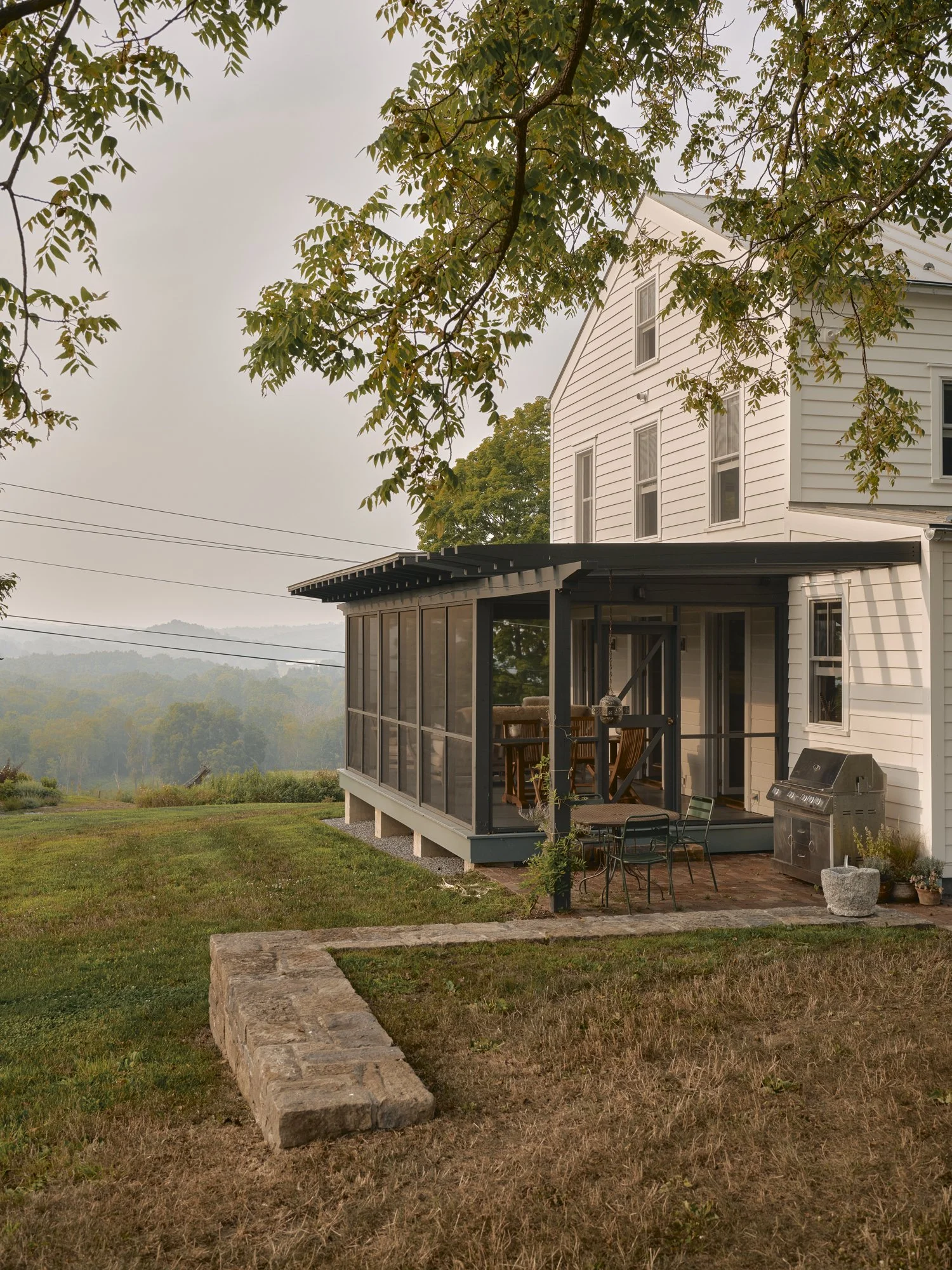 White house with a screened porch, outdoor table and chairs, and a BBQ grill, with a yard and scenic landscape in the background.
