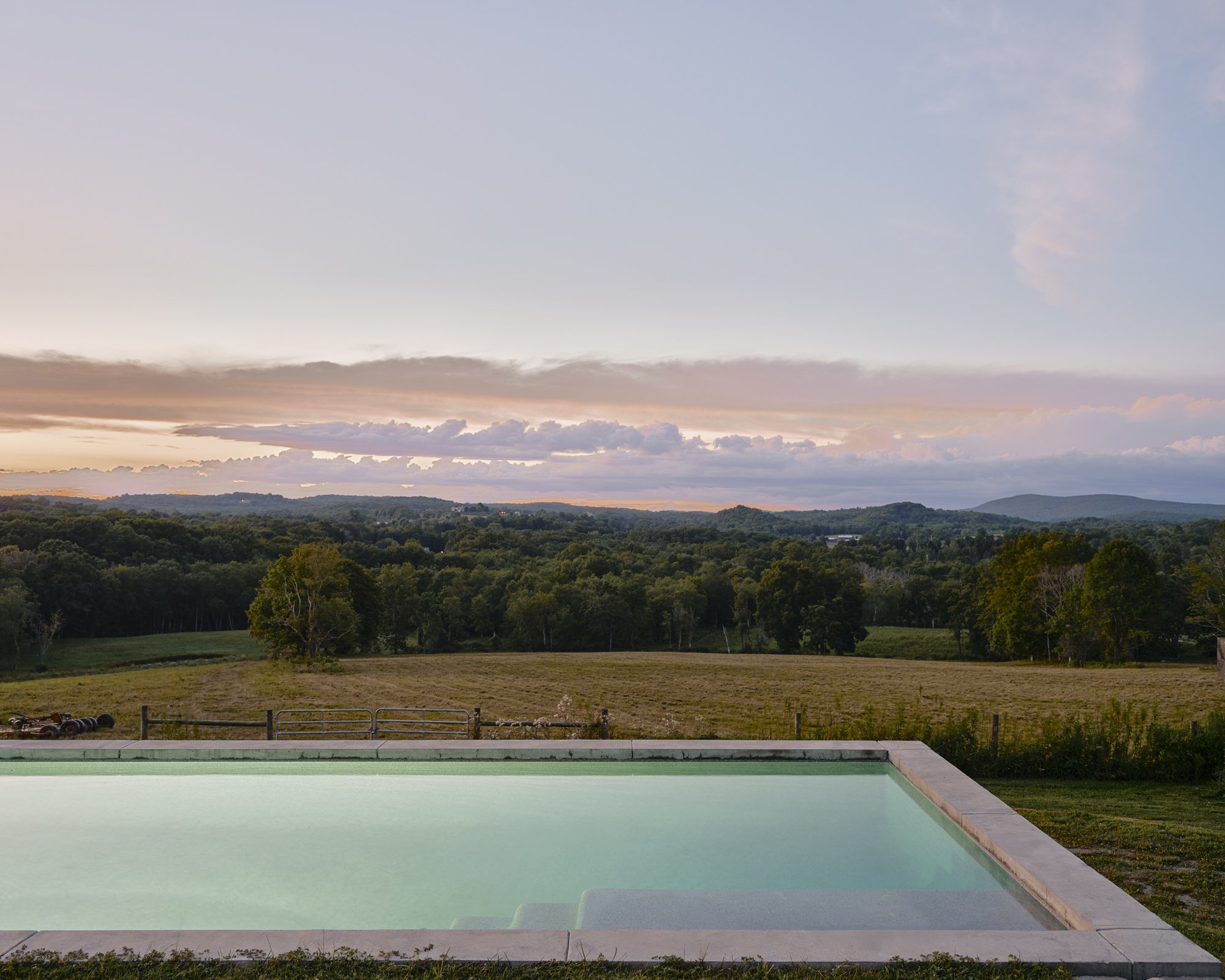 Swimming pool overlooking a rural landscape with fields, trees, and distant hills during sunset.