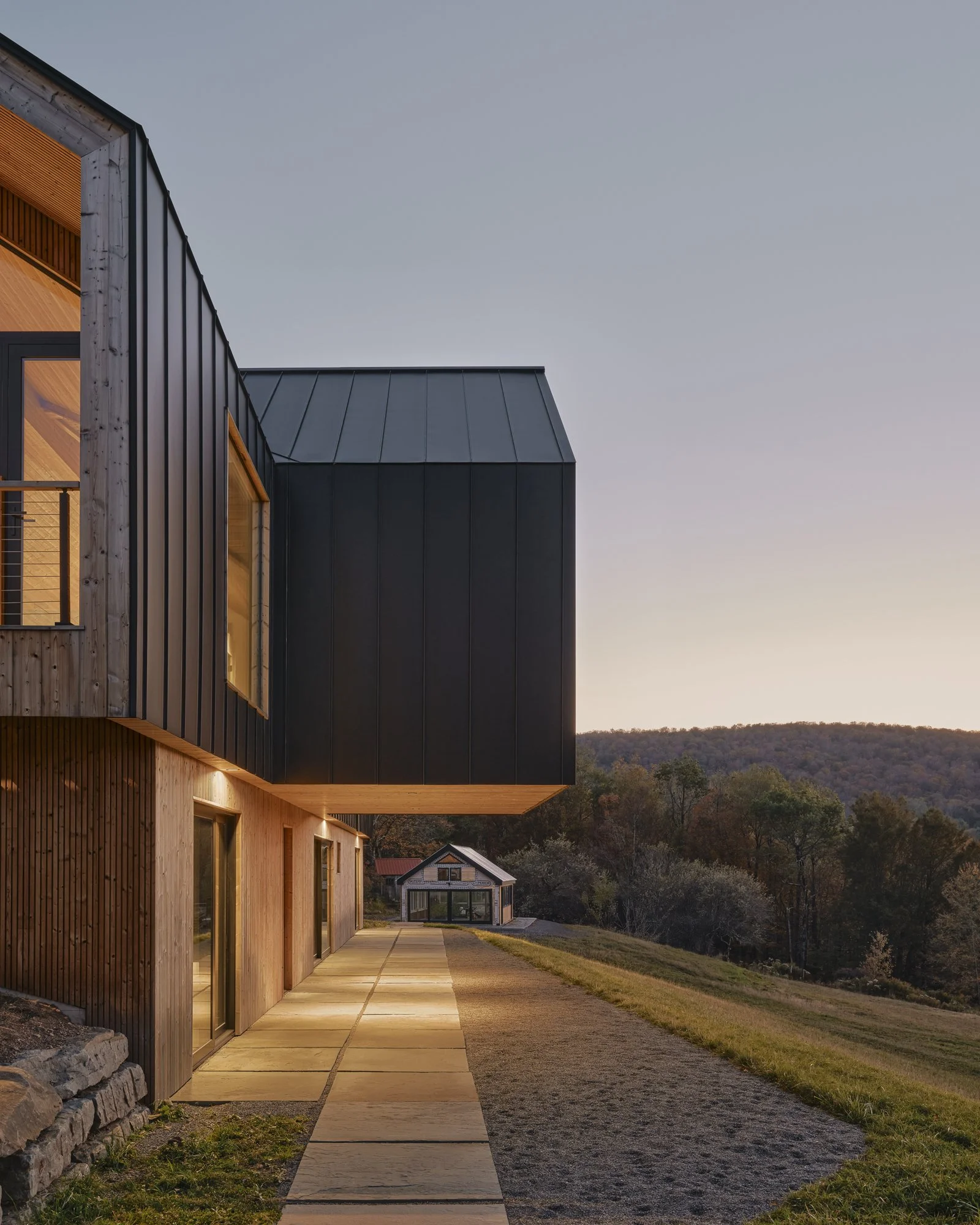 Modern house with black and wooden exterior, illuminated windows, in a rural area with a pathway, trees, and a small building in the distance at sunset.