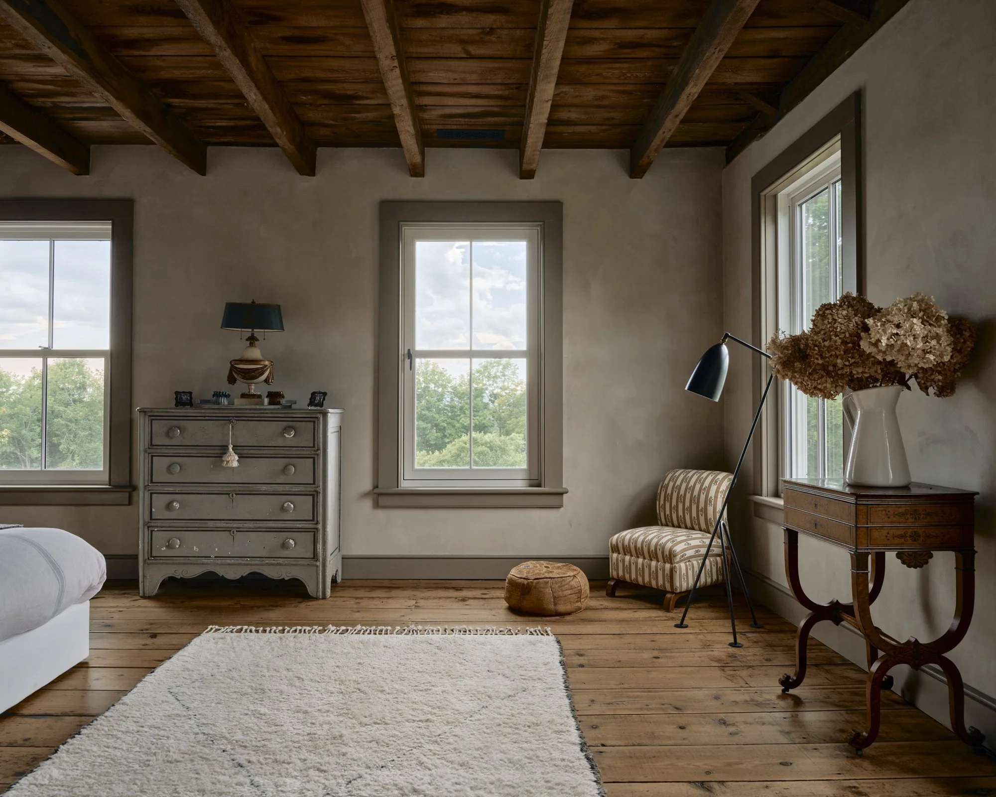 A cozy living room with wooden floors, a cream-colored rug, a white armchair, vintage furniture, a striped armchair, a dried flower arrangement in a white vase, and natural light coming through three windows, with a wood-beamed ceiling.